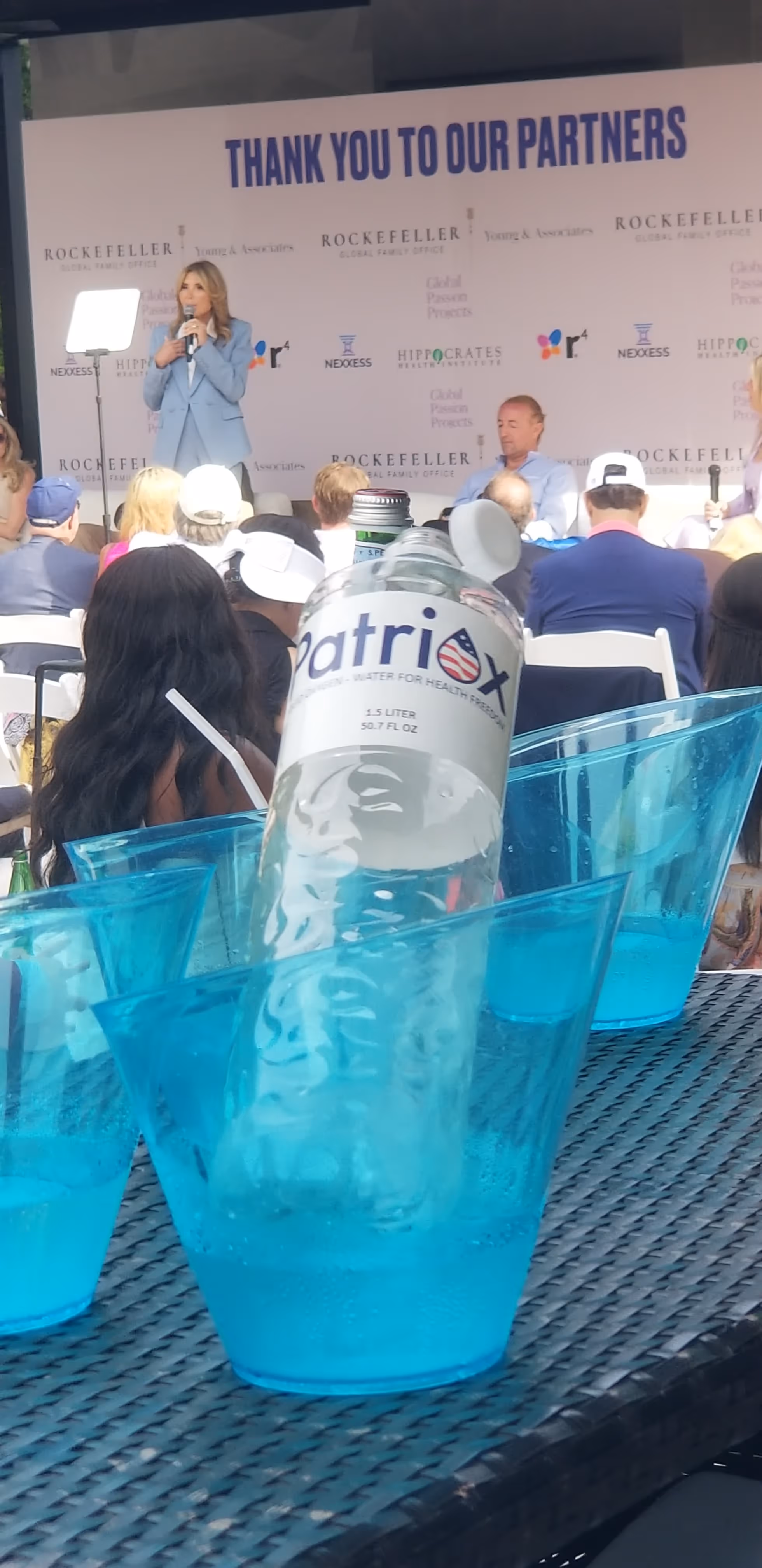 Woman in a blue suit speaking on stage at an event with 'Thank You To Our Partners' banner behind her and people seated in audience; foreground shows a bottle of Patriox water in a blue ice bucket on a table.