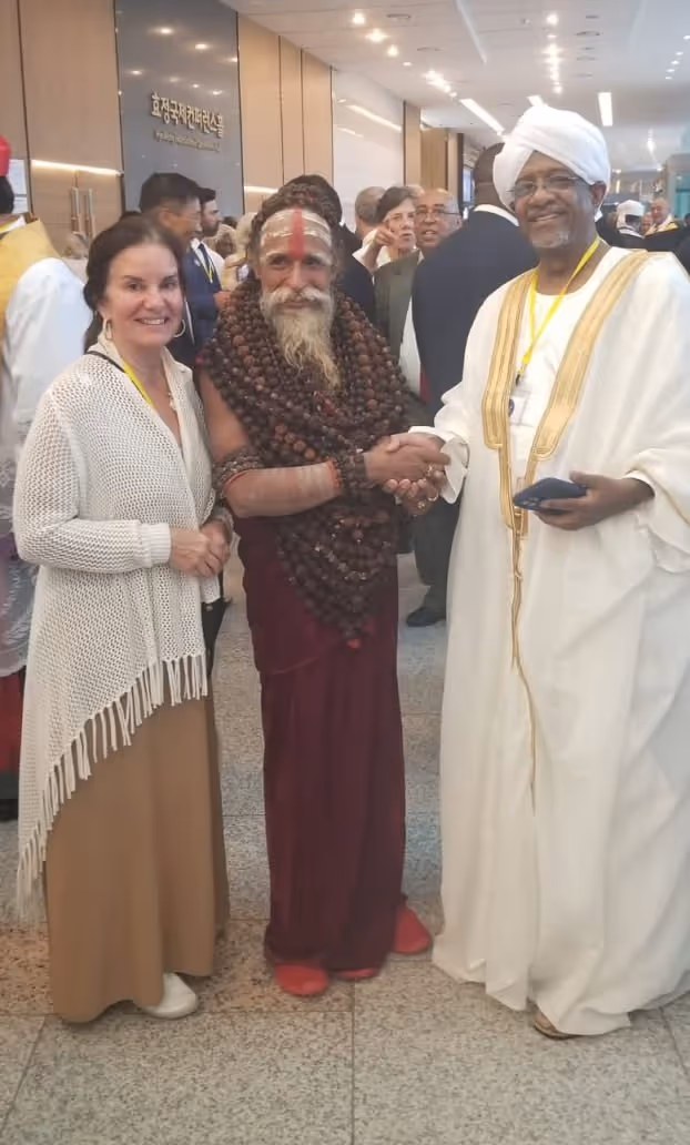 Three people standing indoors at a conference, with a woman on the left smiling, a man in the middle wearing numerous wooden bead necklaces and traditional attire, and a man on the right in white traditional clothing and turban shaking hands with the man in the middle.