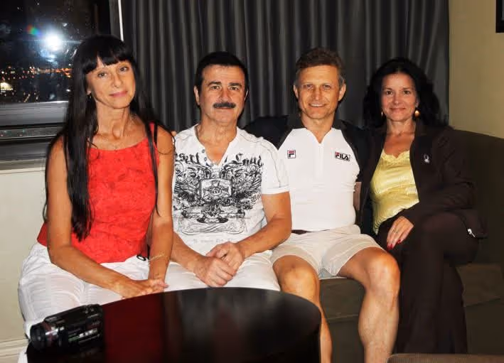 Four adults sitting closely together on a couch indoors, with a camera on a table in front of them and dark curtains in the background.