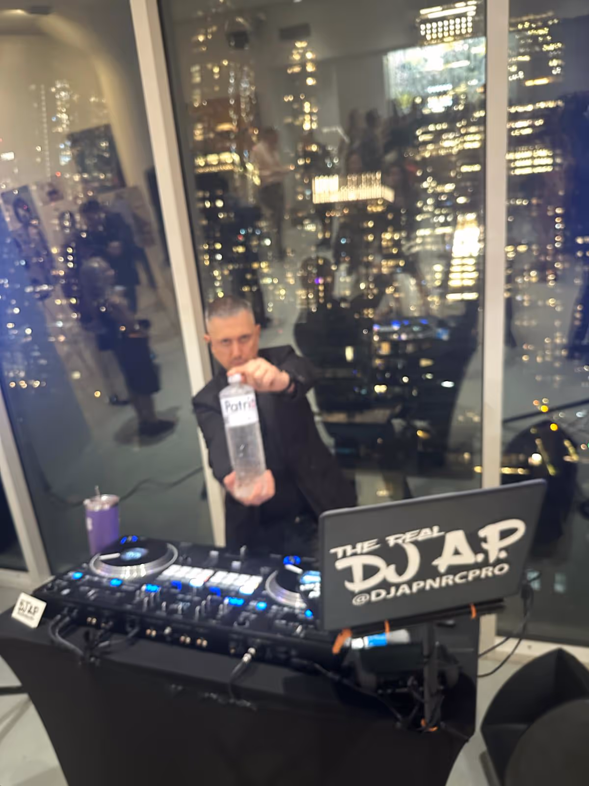 DJ standing behind a DJ booth with a laptop reading 'THE REAL DJ A.P @DJAPNRCPRO,' holding a water bottle labeled 'Patri' in front of a window showing city lights at night.