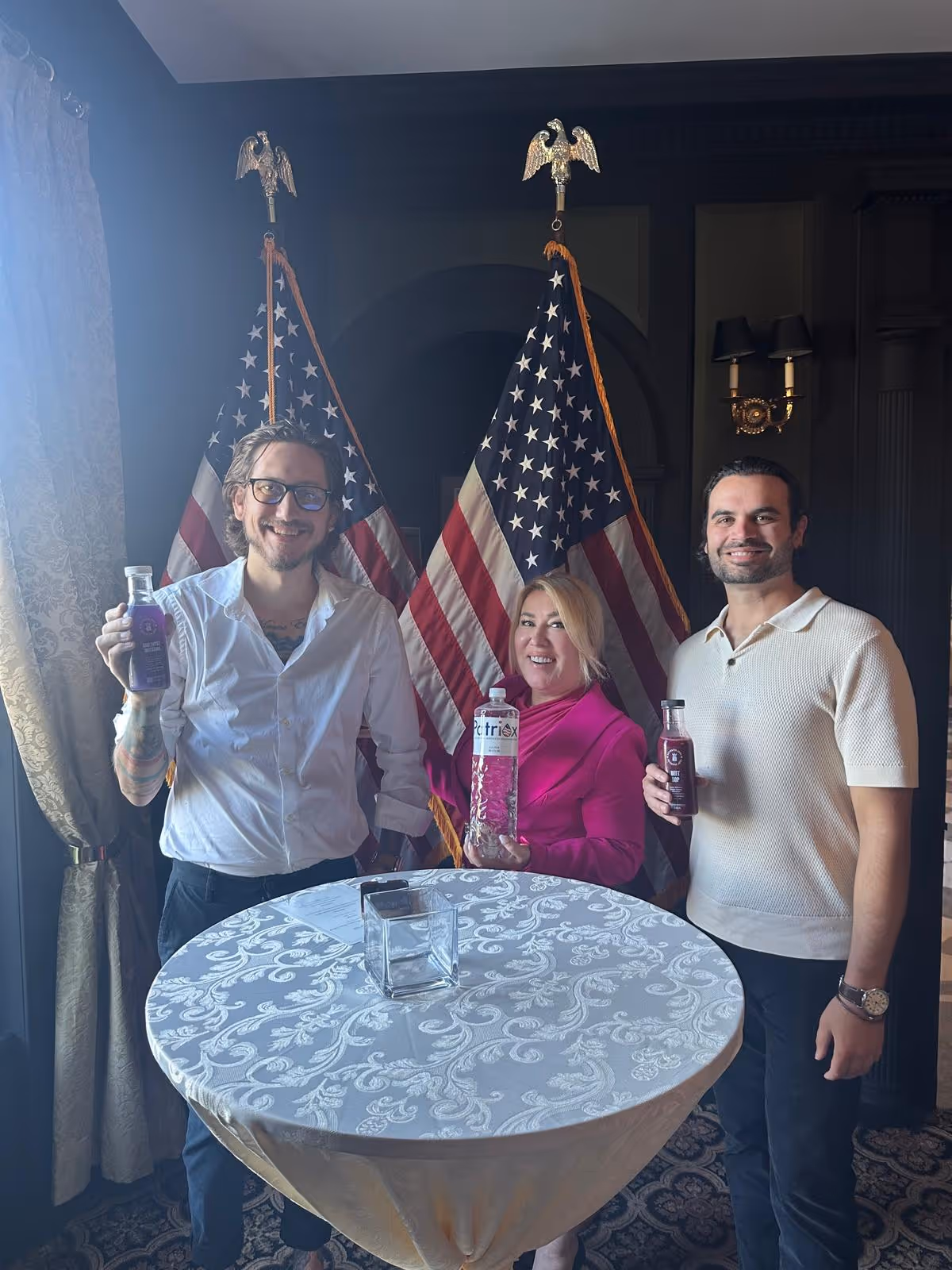 Three people standing behind a round table holding bottled beverages with two American flags in the background.