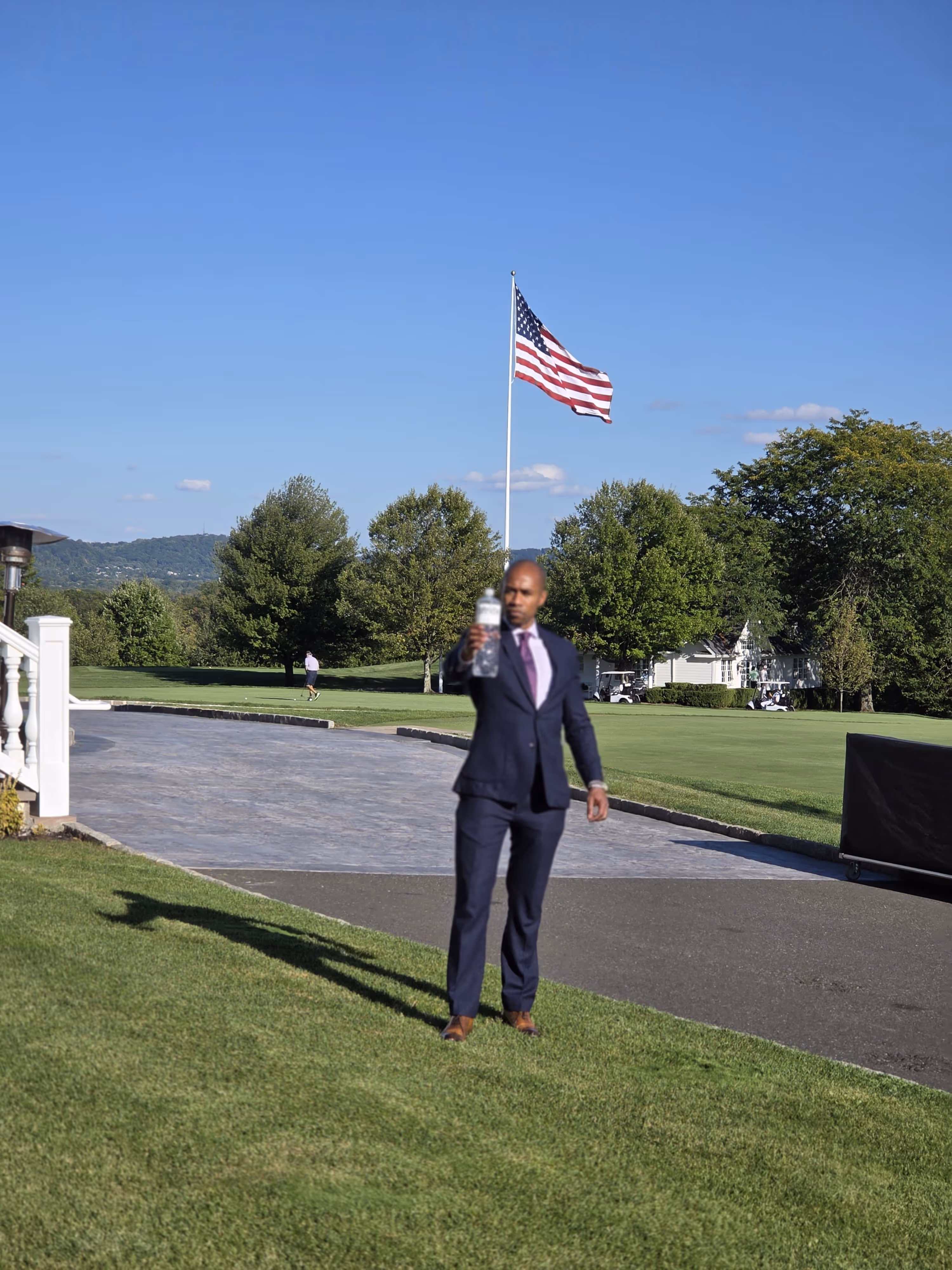 Man in a dark suit holding a water bottle outdoors with an American flag and trees in the background on a sunny day.
