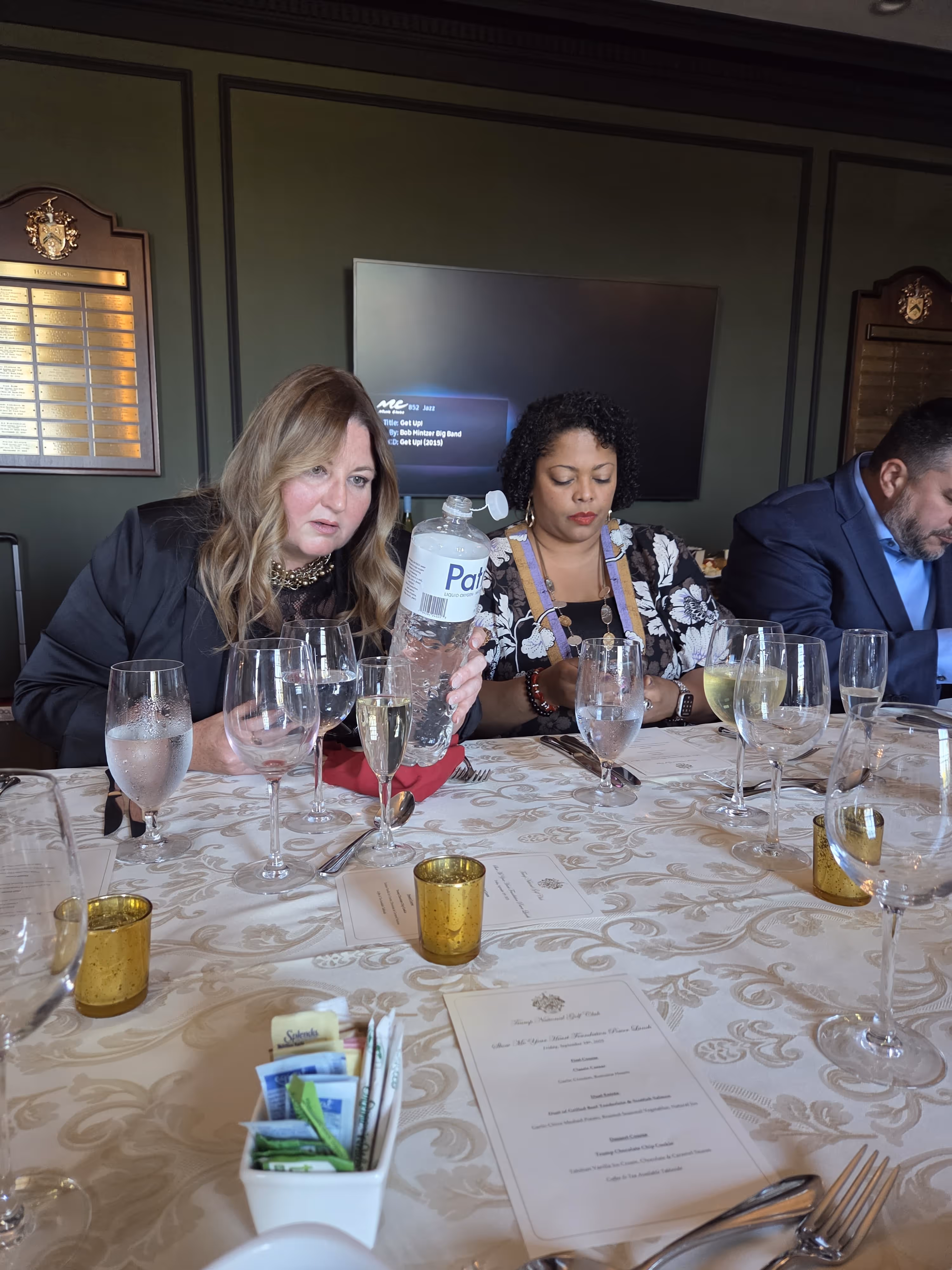 Three people sitting at a table with tablecloth, menus, multiple glasses, and a bottle of water in a formal dining setting.