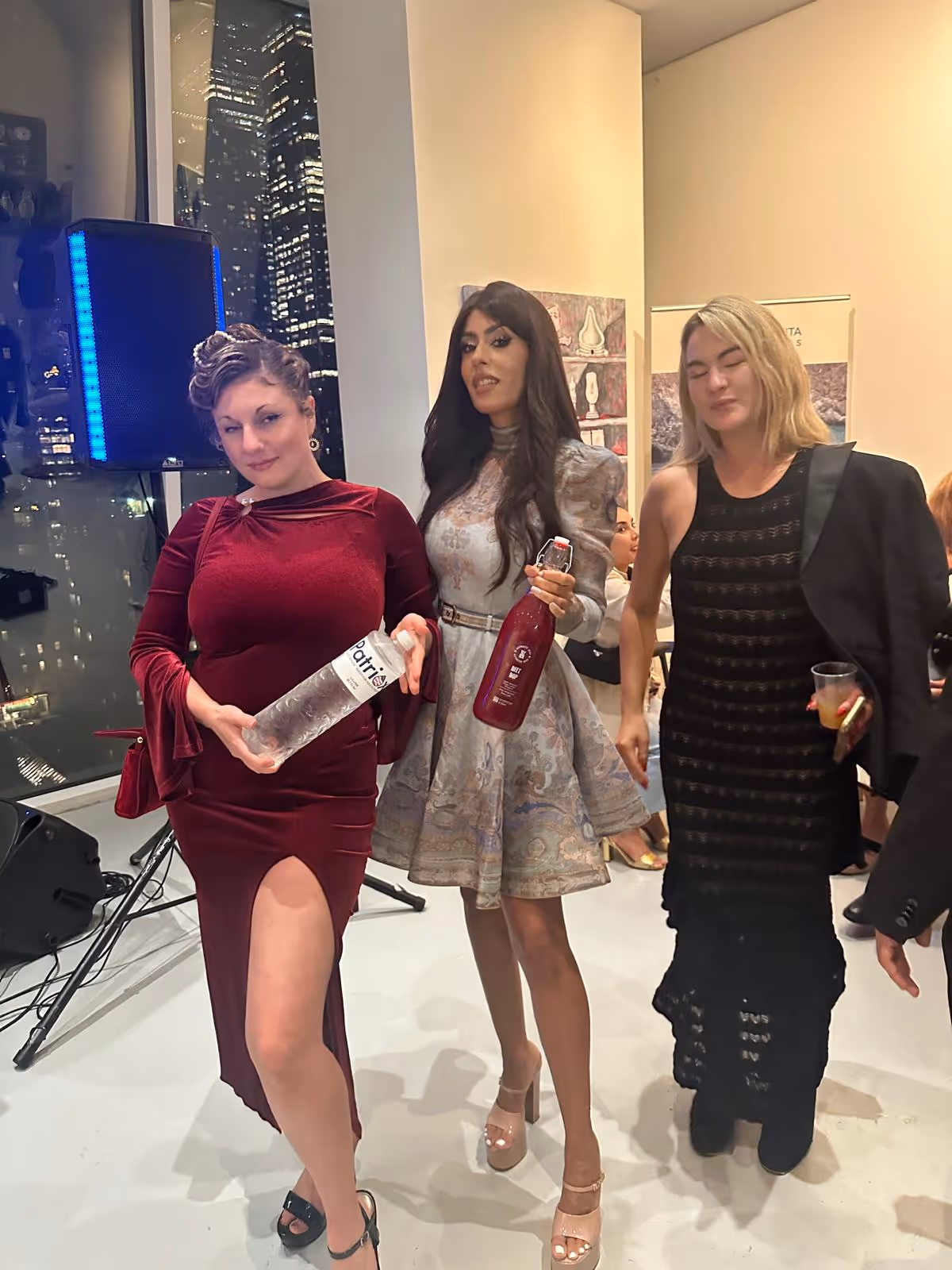 Three women dressed elegantly at an indoor event, two holding bottles and one holding a drink, with city lights visible through a large window behind them.
