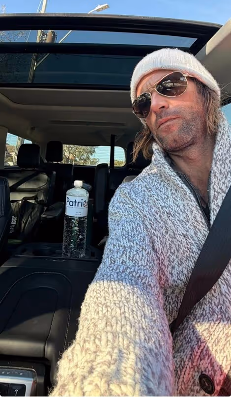 Man wearing sunglasses and white knit hat taking a selfie inside a car with sunroof open and a water bottle labeled 'Patri' on the center console.