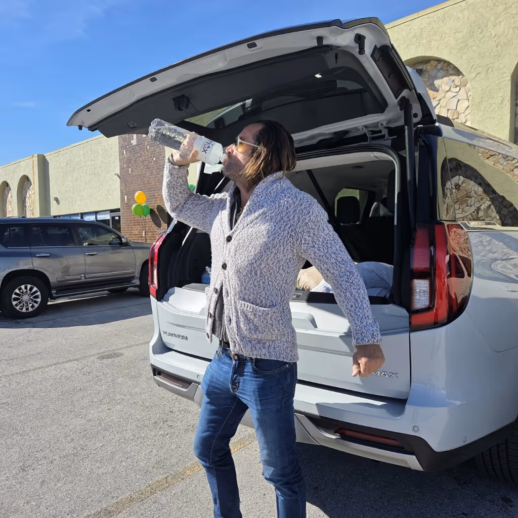 Man wearing a gray sweater and sunglasses drinking from a bottle while standing next to an open white SUV trunk in a sunny parking lot.