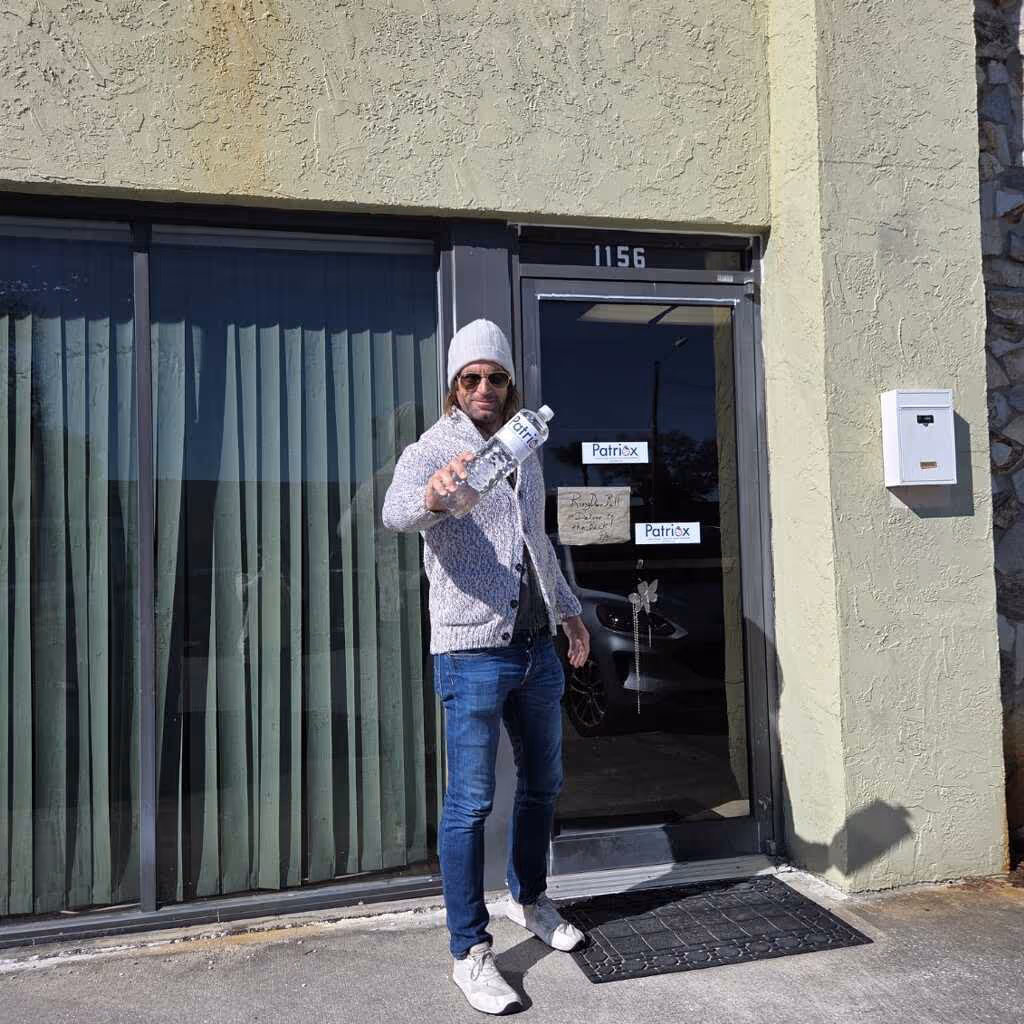 Man wearing a beanie, sunglasses, and gray sweater standing outside a building with green curtains, holding a bottle of water labeled PatriX.