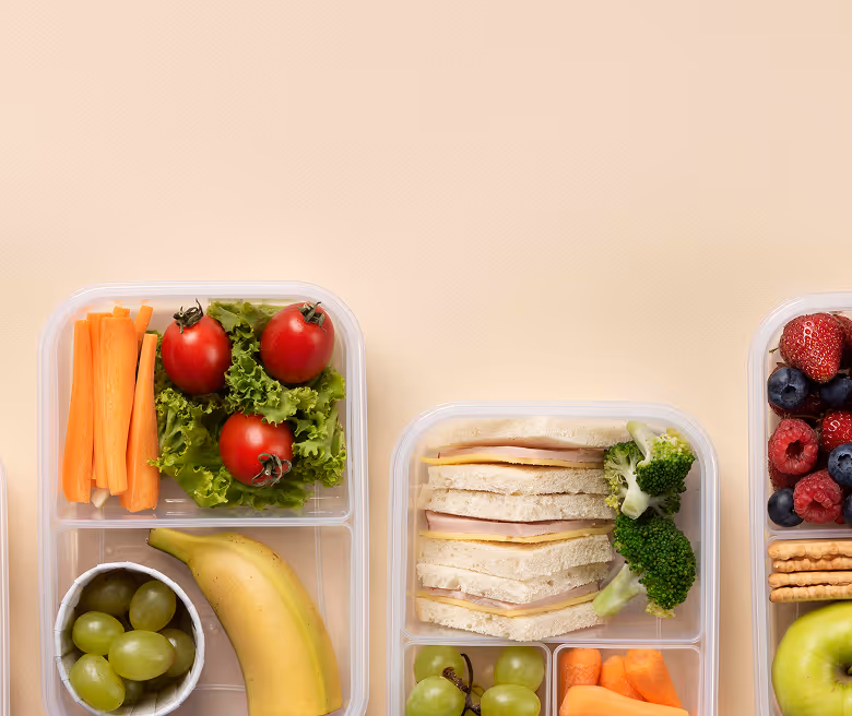 Three compartment meal prep containers with healthy food including carrot sticks, cherry tomatoes on lettuce, grapes, a banana, sandwich slices, broccoli, mixed berries, and crackers.