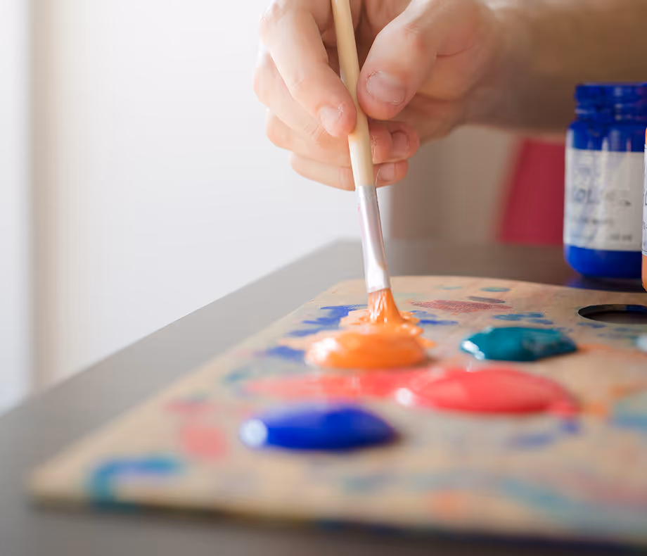 Hand holding a paintbrush dipping into orange paint on a palette with various paint colors.
