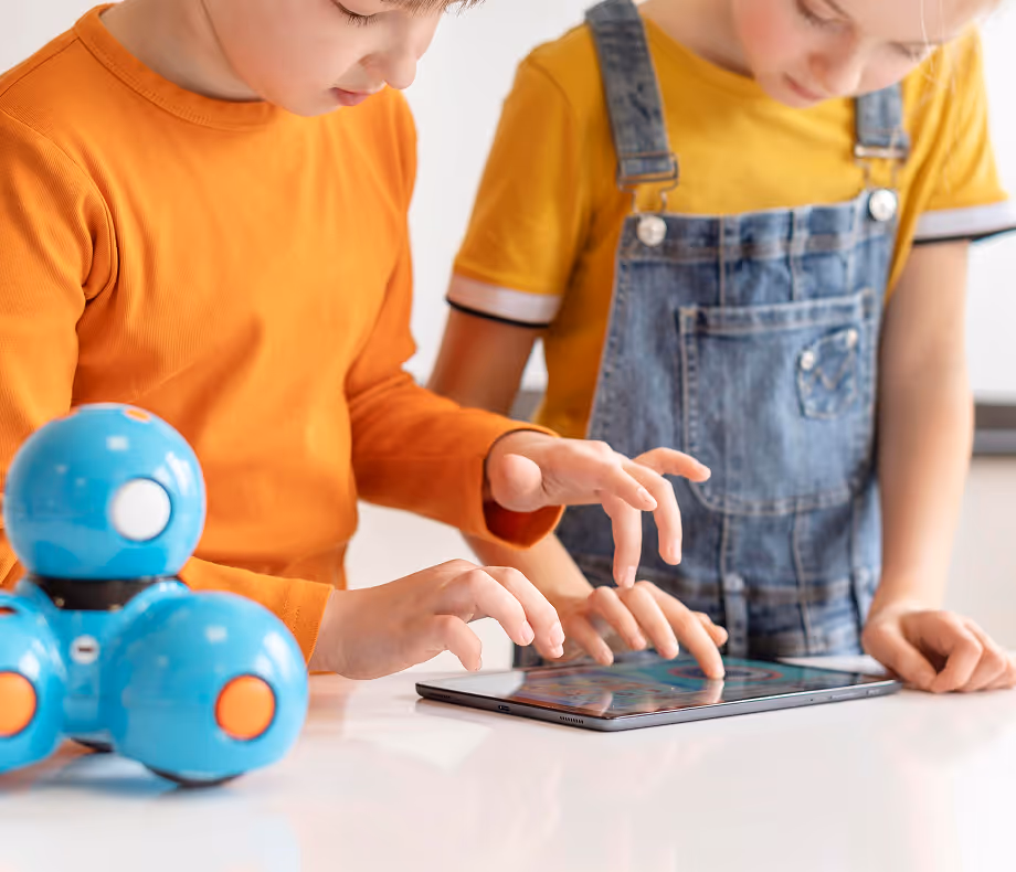 Two children using a tablet together with a blue educational robot on the table nearby.