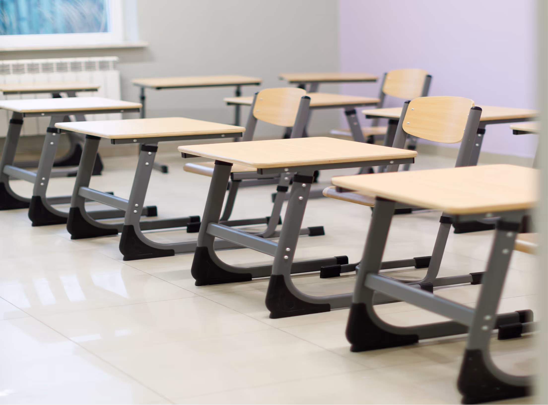 Row of empty school desks and chairs in a bright classroom with tiled floor and large window.