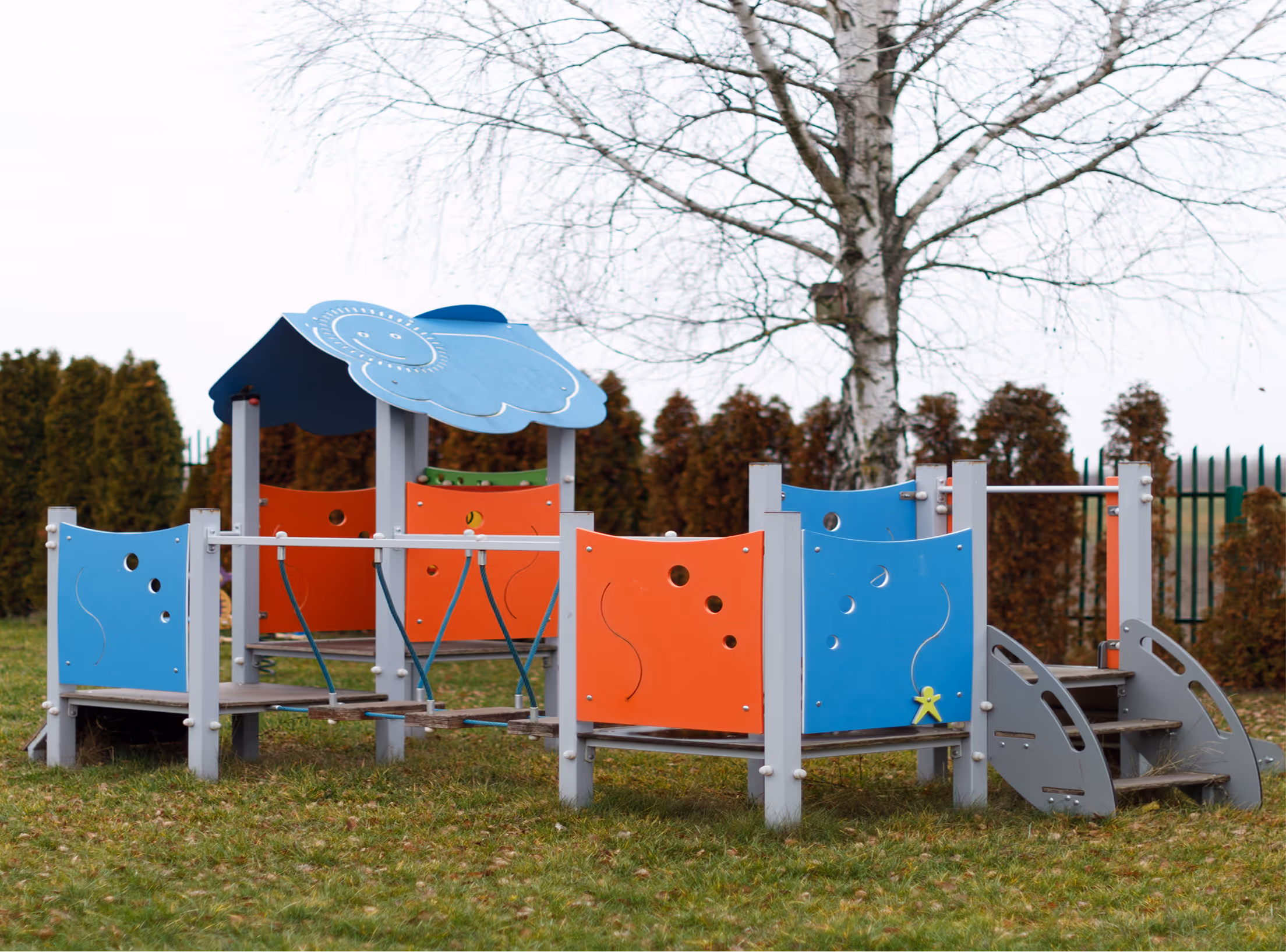 Colorful playground structure with blue and orange panels, wooden steps, and a roof shaped like a cloud on green grass.