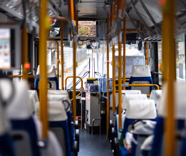 Interior of a bus with blue and white seats, yellow handrails, and a driver wearing a face mask visible in the rearview mirror.