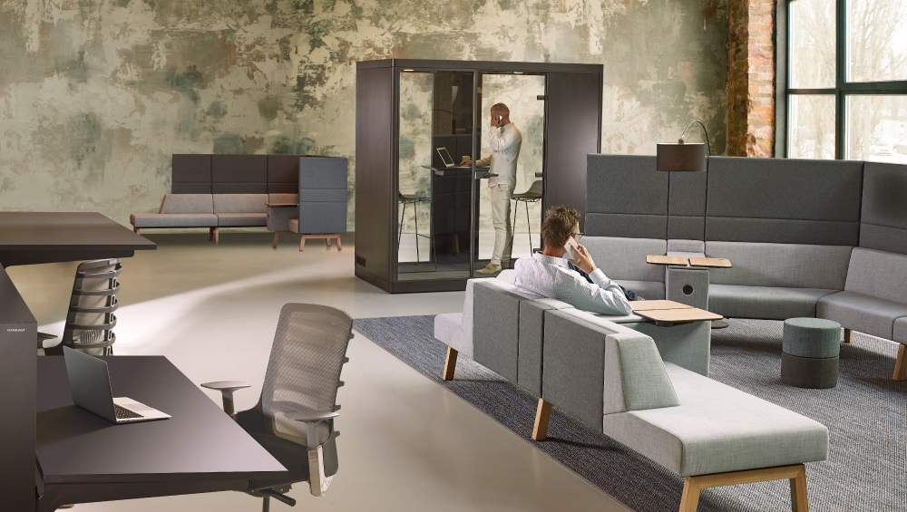 Modern kantoor met grijze modulaire banken, een telefooncabine en een persoon zittend terwijl die telefoneert.