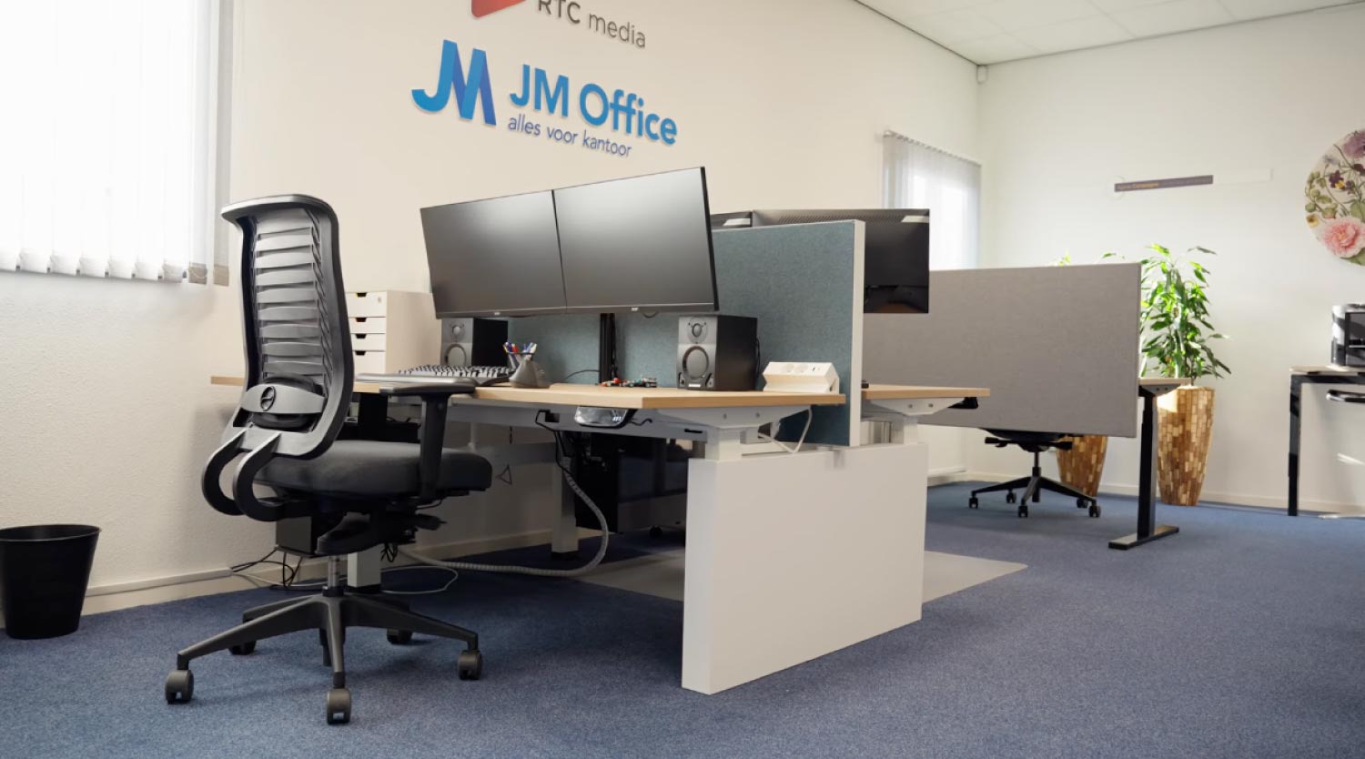 Modern kantoorwerkplek met ergonomische stoel, twee beeldschermen, speakers en bureauaccessoires, tegen een muur met het JM Office-logo.