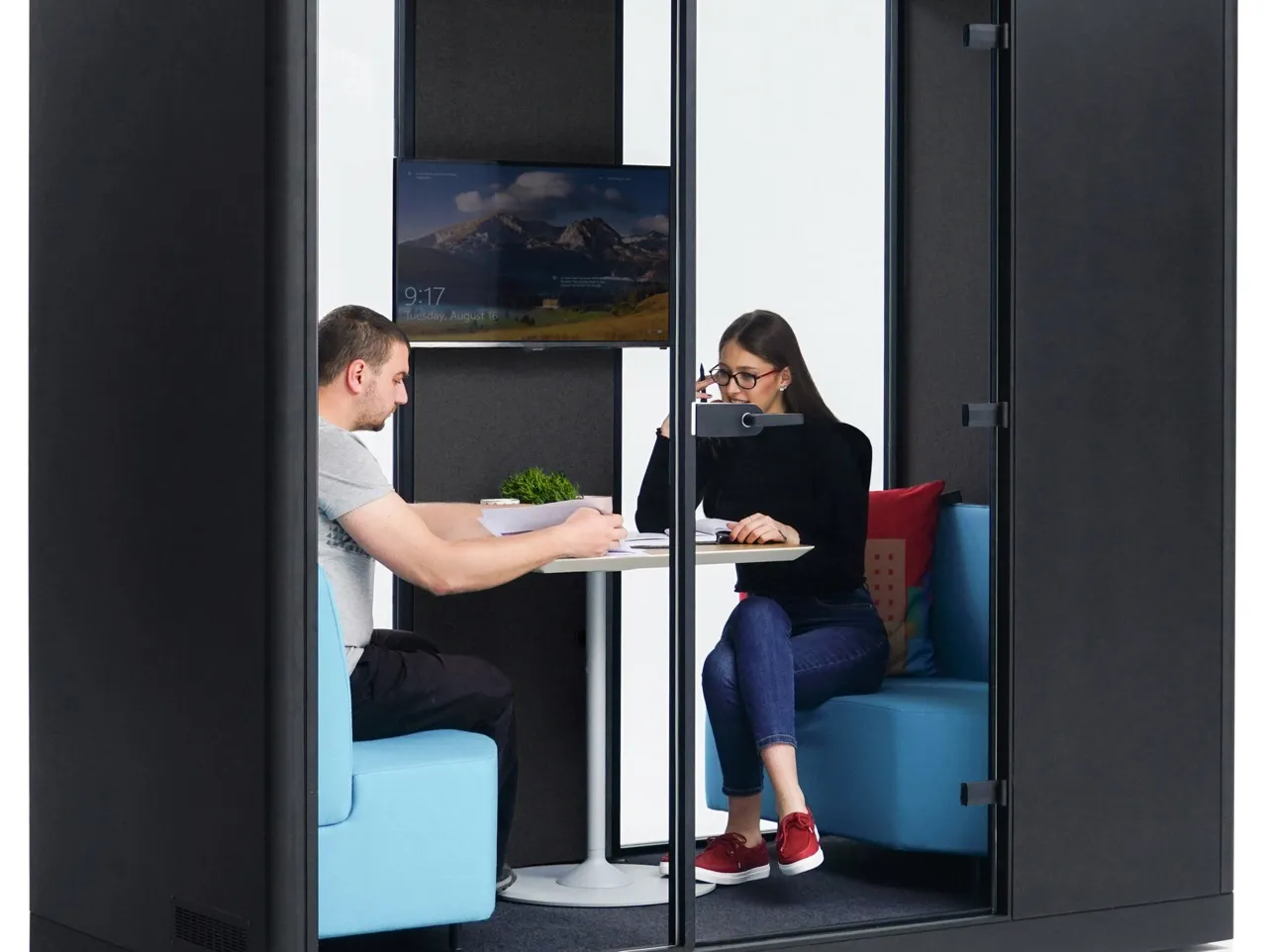 Twee mensen zittend in een moderne kantoortelefooncel met blauwe zitplaatsen, een klein rond tafeltje en een scherm aan de muur.
