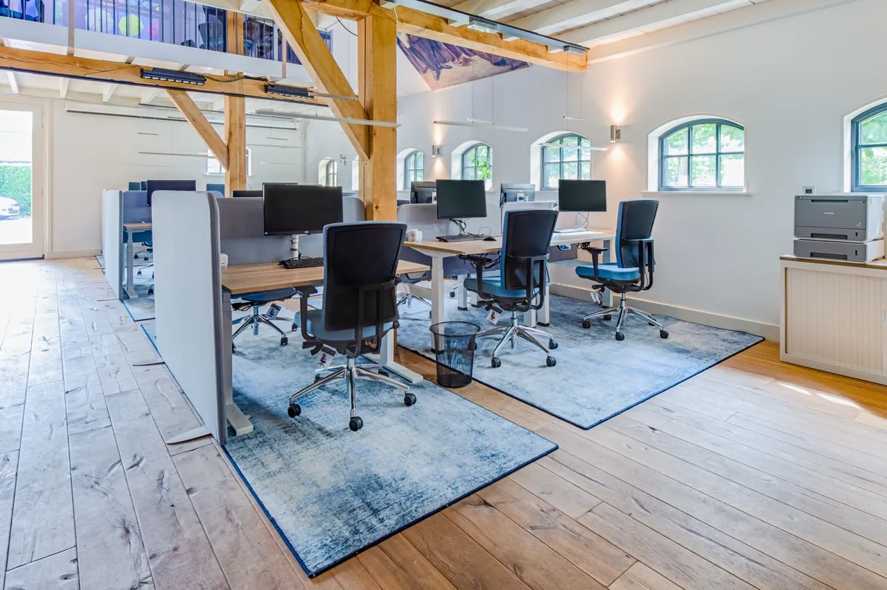 Licht modern kantoor met houten vloeren, werkplekken gescheiden door grijze scheidingswanden, zwarte bureaustoelen en computers op bureaus met blauwe vloerkleden eronder.
