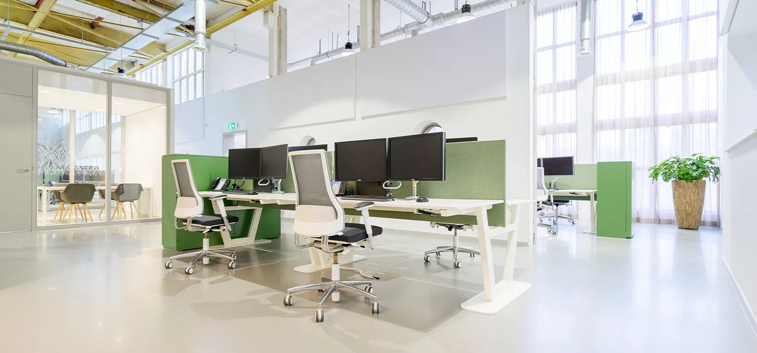 Modern open kantoor met witte bureaus, ergonomische stoelen, computerschermen, groene privacypanelen en grote ramen met doorschijnende gordijnen.
