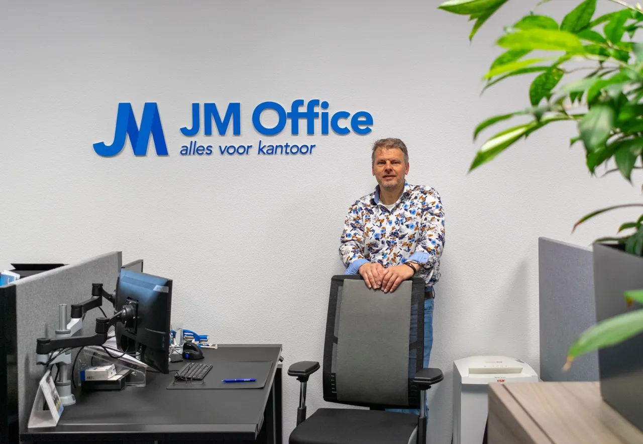 Jan Marcel staand achter een bureaustoel in een modern kantoor, met het JM Office-logo en de slogan op de muur.
