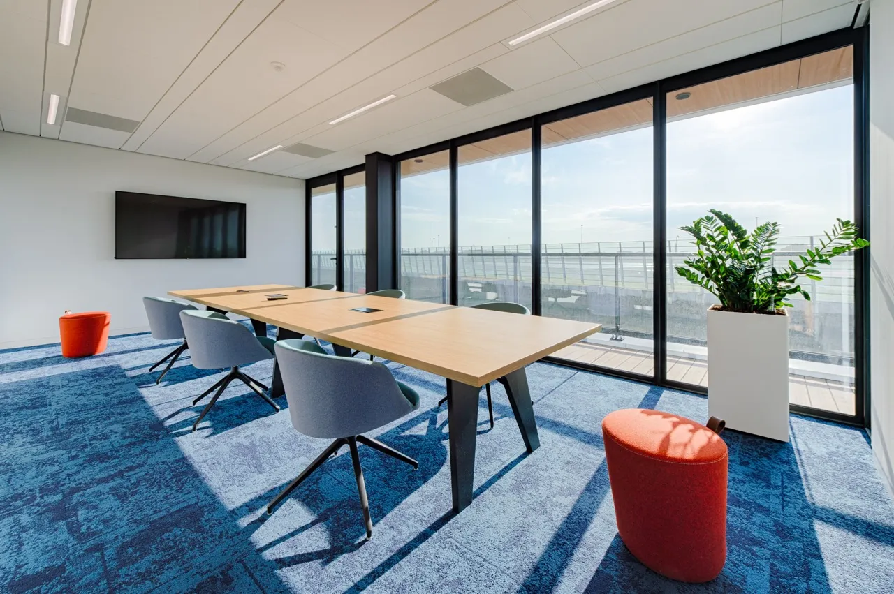 Moderne kantoorvergaderruimte met een lange houten tafel, blauwe stoelen, grote ramen van vloer tot plafond, een blauw tapijt, twee oranje poefs en een groene kamerplant.
