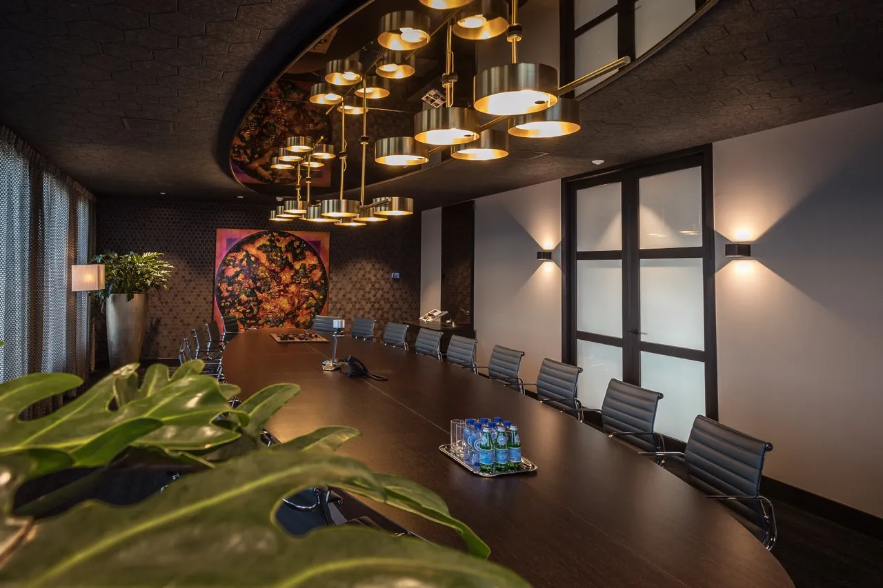 Moderne vergaderruimte met een lange tafel van donker hout, zwarte leren stoelen, een grote groene plant en eigentijdse hanglampen.
