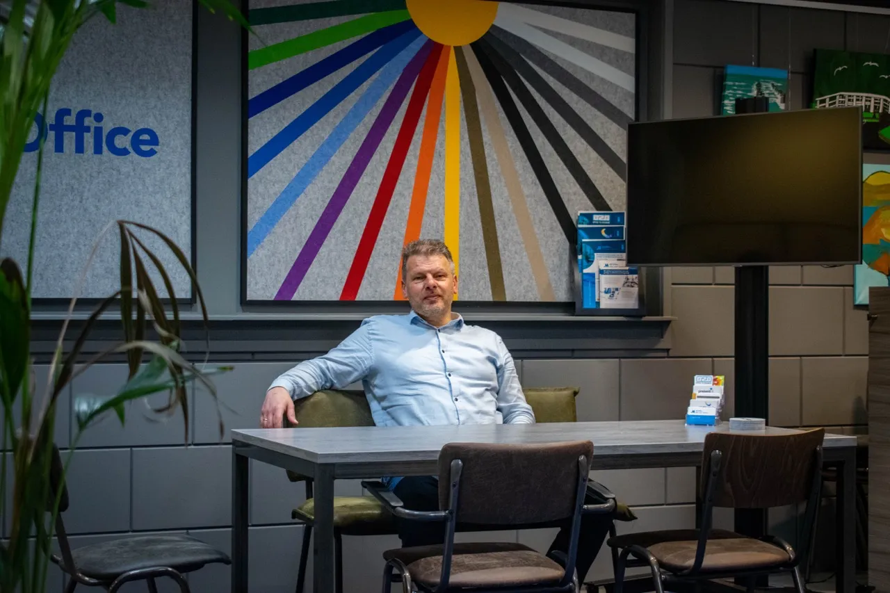 Jan Marcel in een lichtblauw shirt, zittend aan een tafel in een modern kantoor met kleurrijke abstracte wandkunst en een groot beeldscherm.
