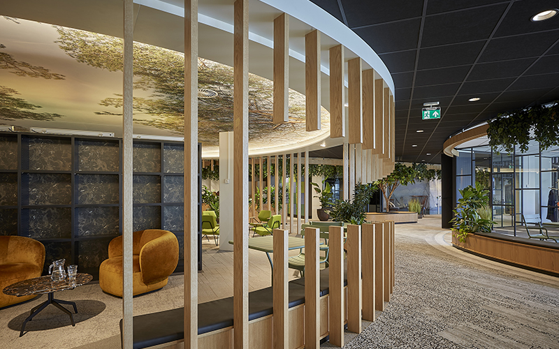 Moderne binnenlounge met houten lattenwanden, mosterdgele fauteuils, groene stoelen, planten en een gebogen plafondlamp.