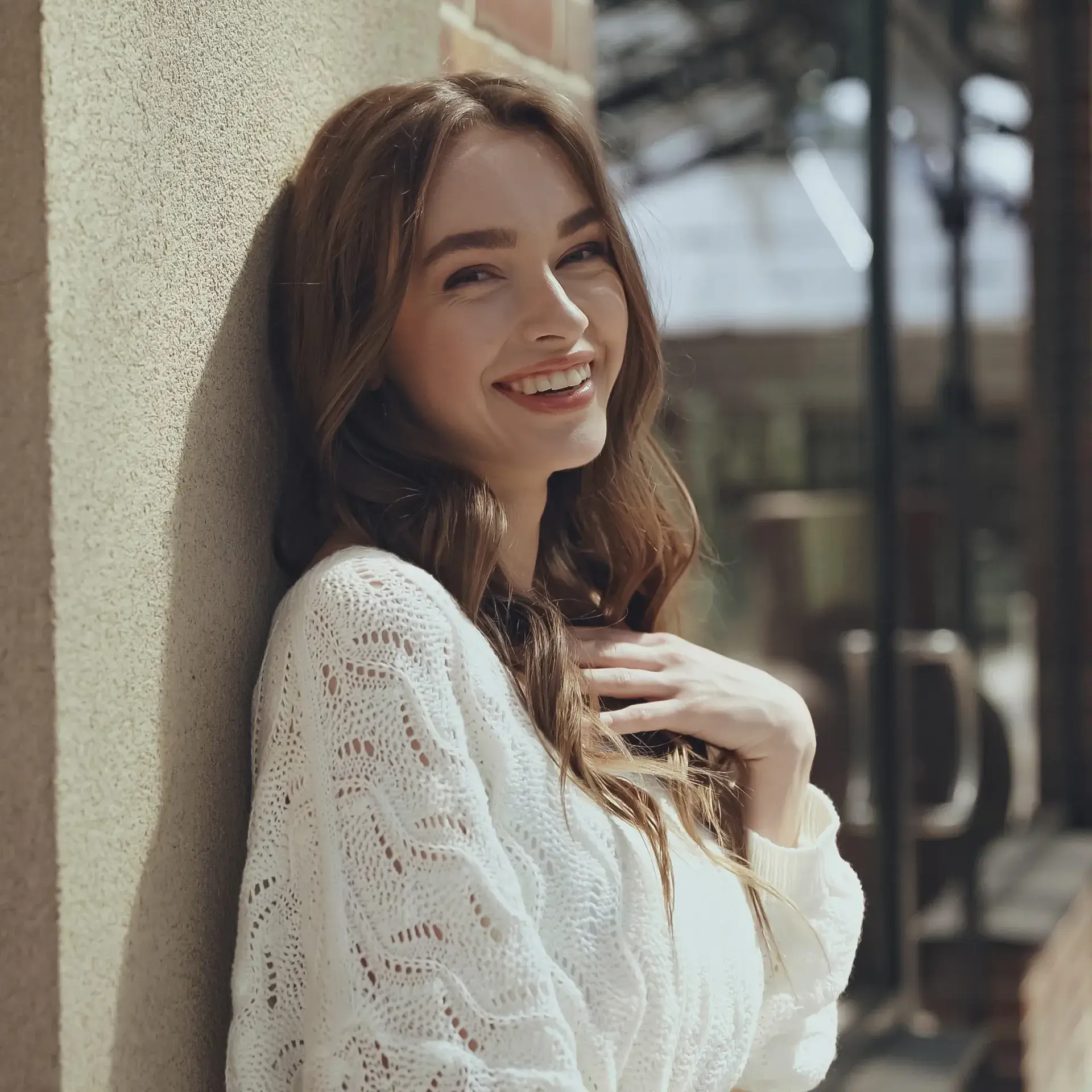 Smiling woman with long brown hair wearing a white knitted sweater leaning against a beige wall.
