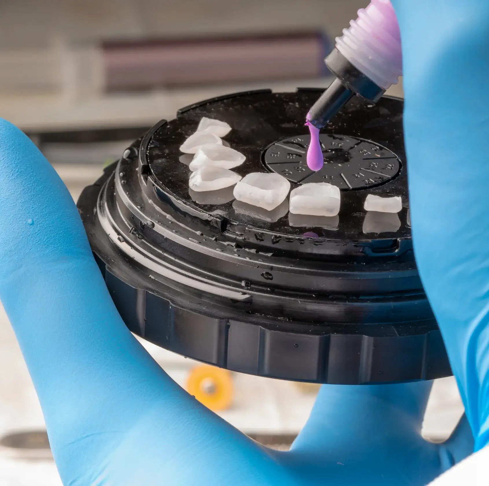 Gloved hand holding a black palette with dental veneers and applying purple liquid from a small bottle onto one veneer.