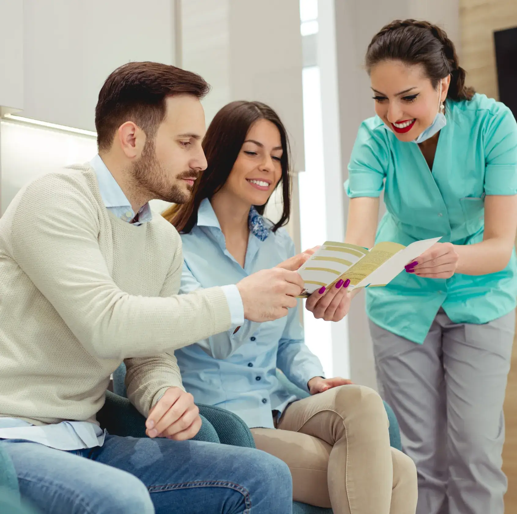 Dentist in teal scrubs showing a brochure to a smiling couple seated in a waiting area.