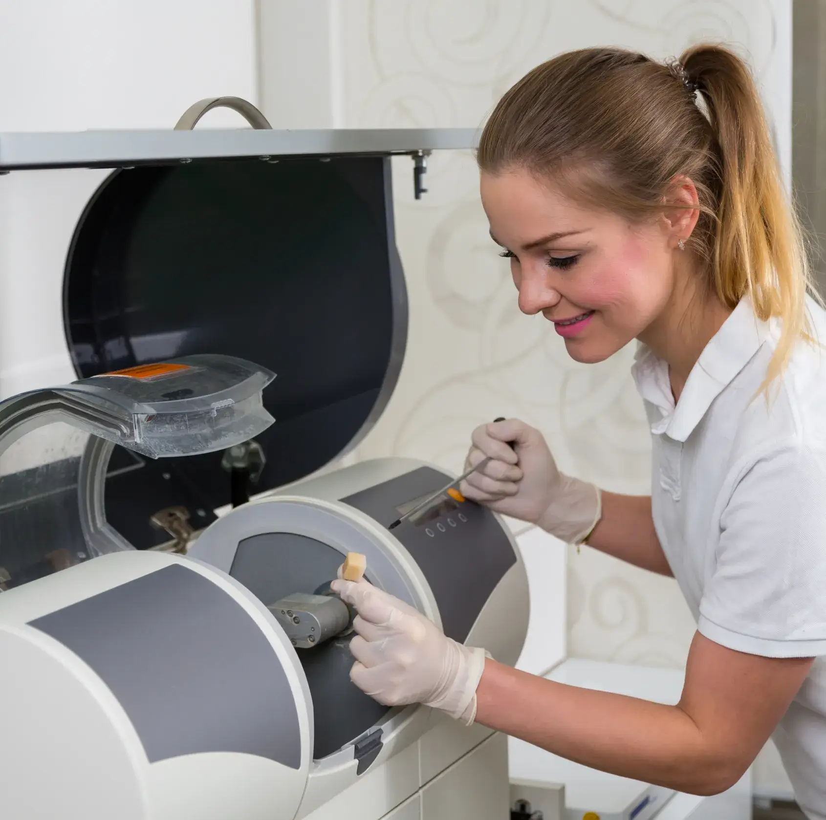 Smiling woman wearing gloves operating a dental milling machine with a dental crown mold.