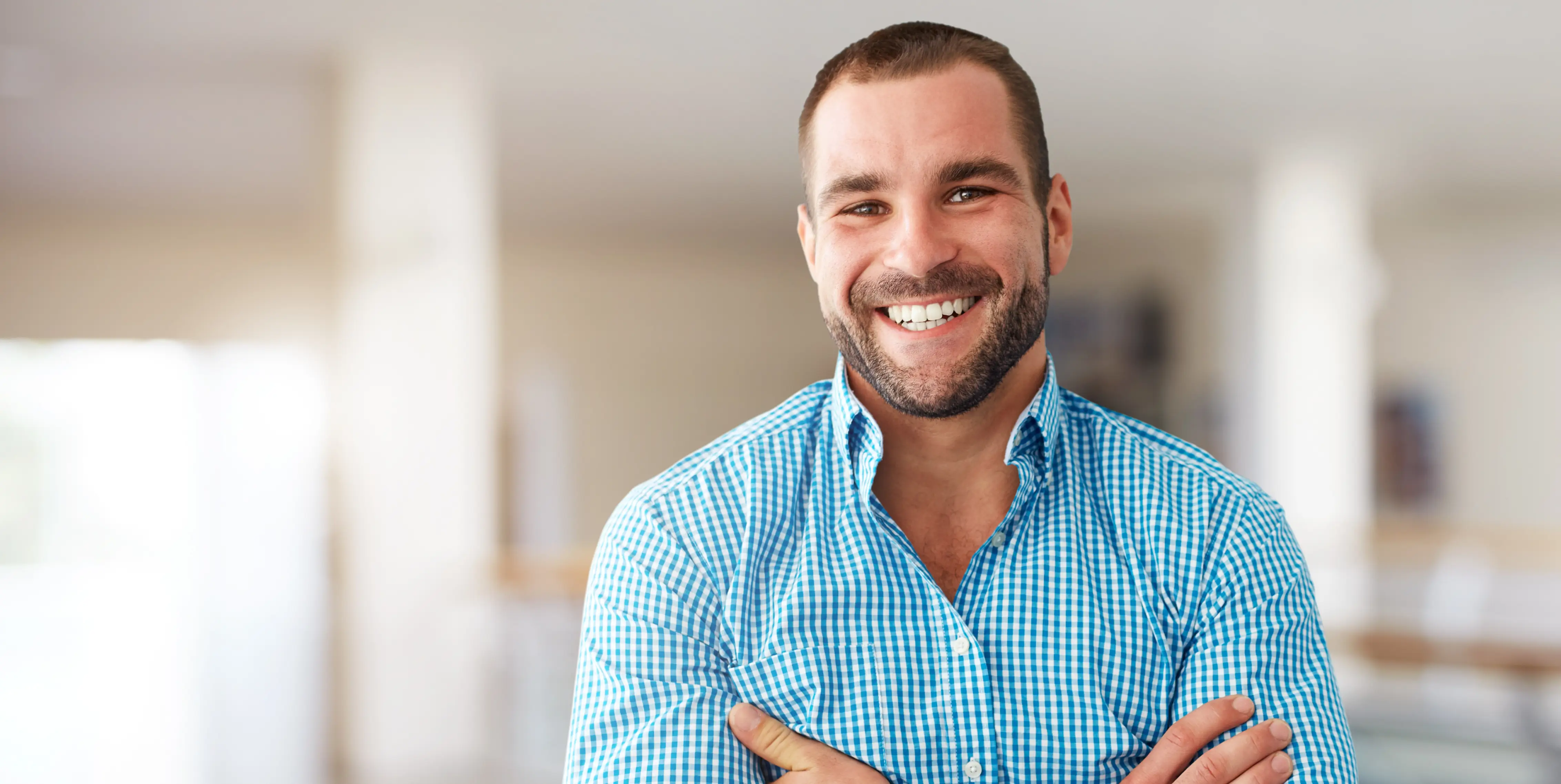 Smiling man with short hair and beard wearing a blue checkered shirt, standing with arms crossed in a bright room.