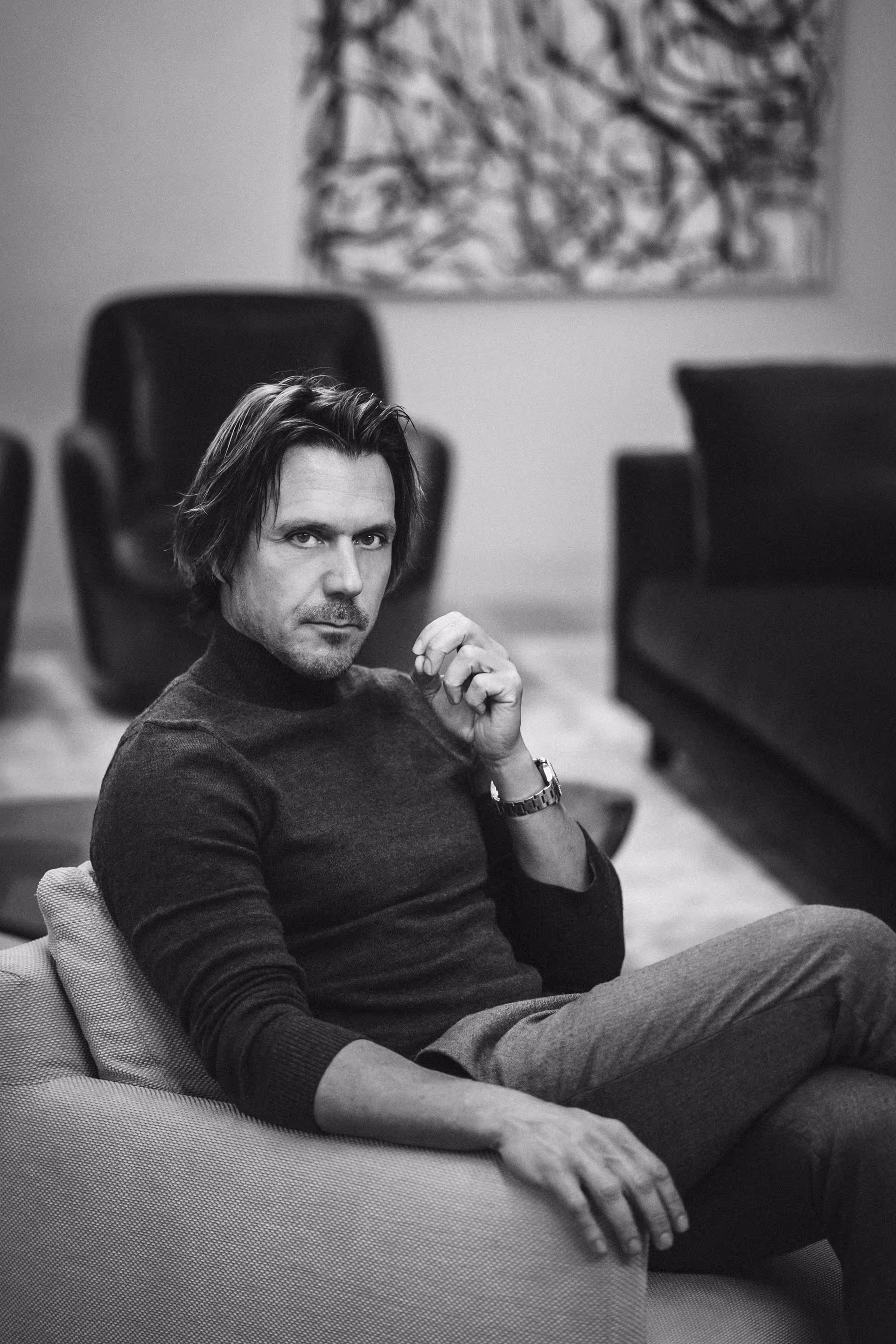 Black and white photo of a man with medium-length hair wearing a dark turtleneck sweater, sitting on a couch with a serious expression.