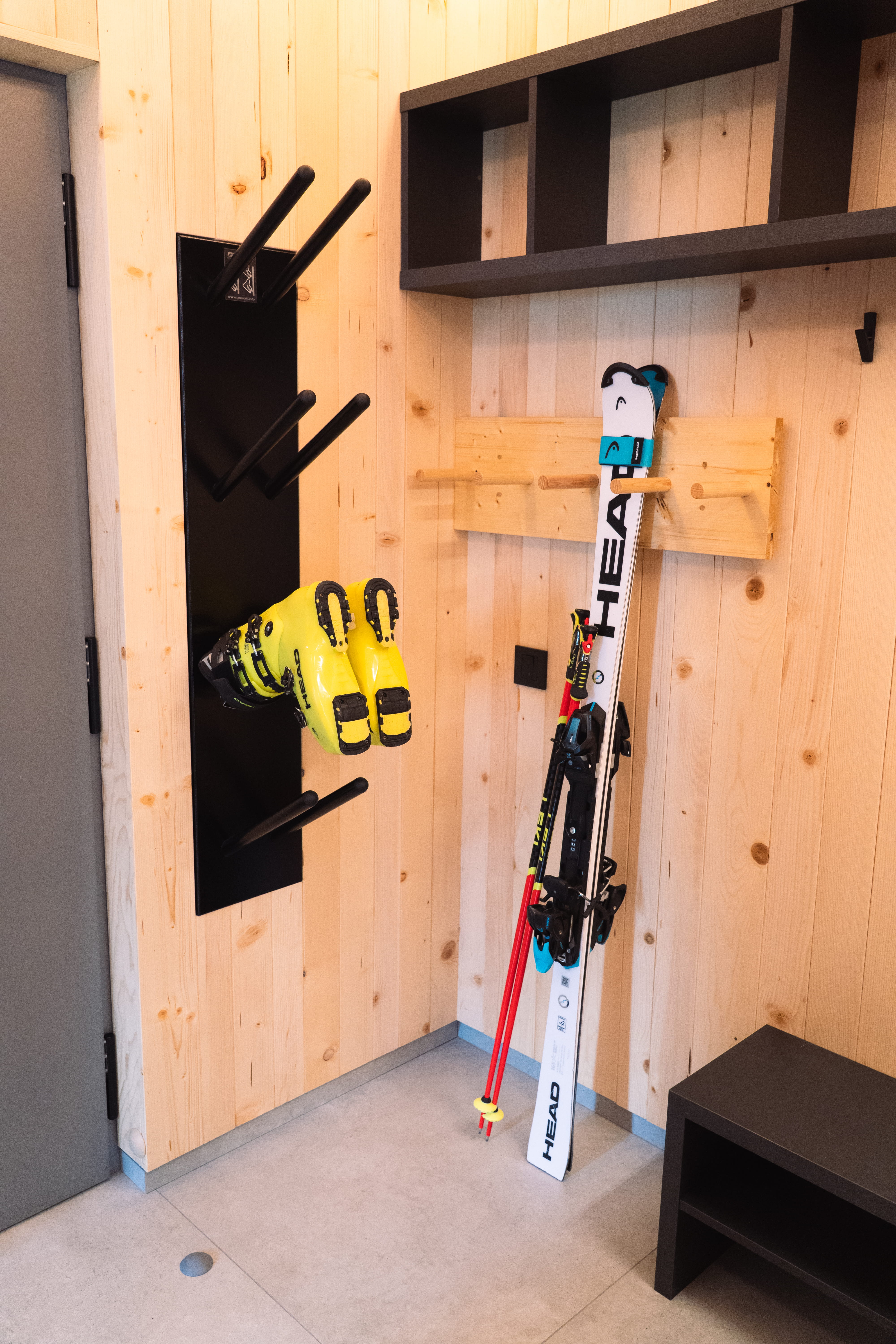 Ski boots hanging on a black wall rack and skis with poles leaning against a light wooden paneled wall.