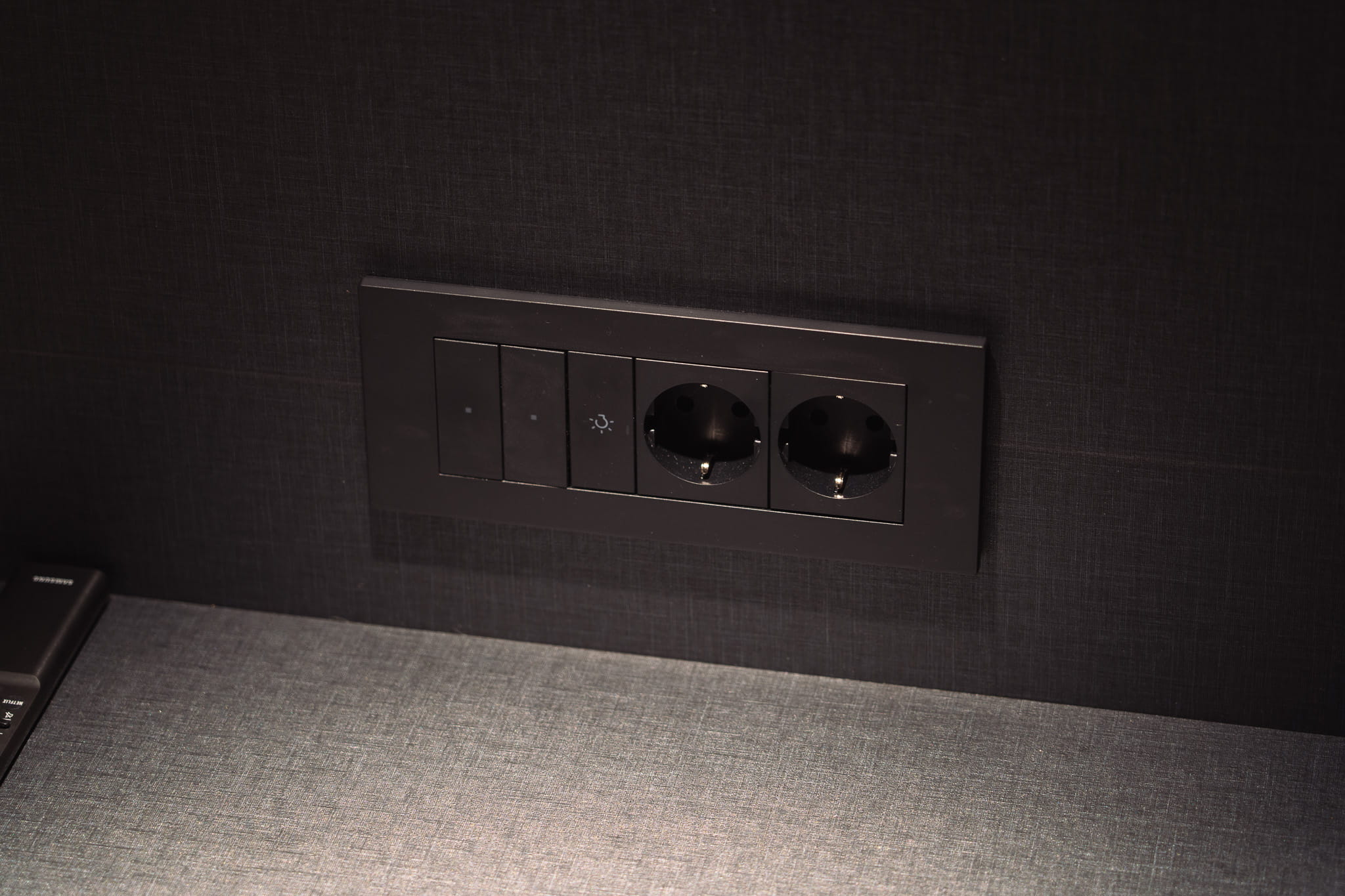Black wall panel with three light switches and two European power outlets above a gray surface.