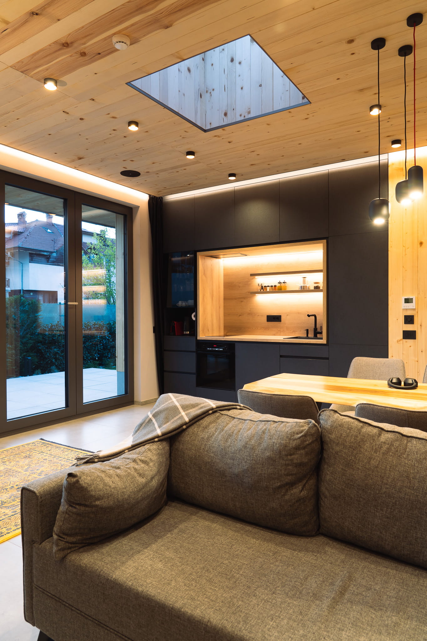 Modern living room with gray sofa, wooden ceiling, skylight, and small kitchen area with black cabinets.