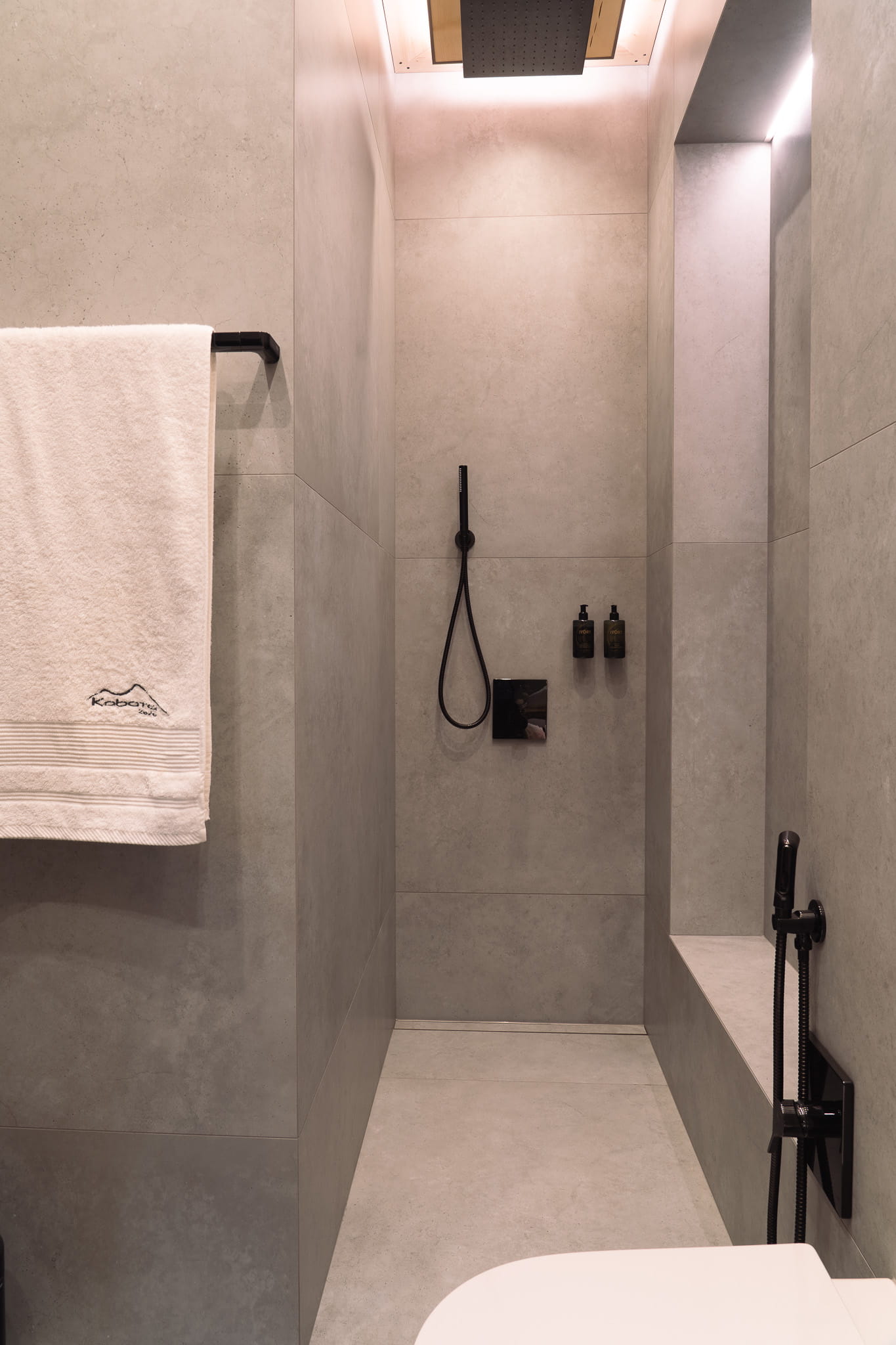 Minimalist bathroom shower area with gray tiles, black fixtures and white towel on rack.