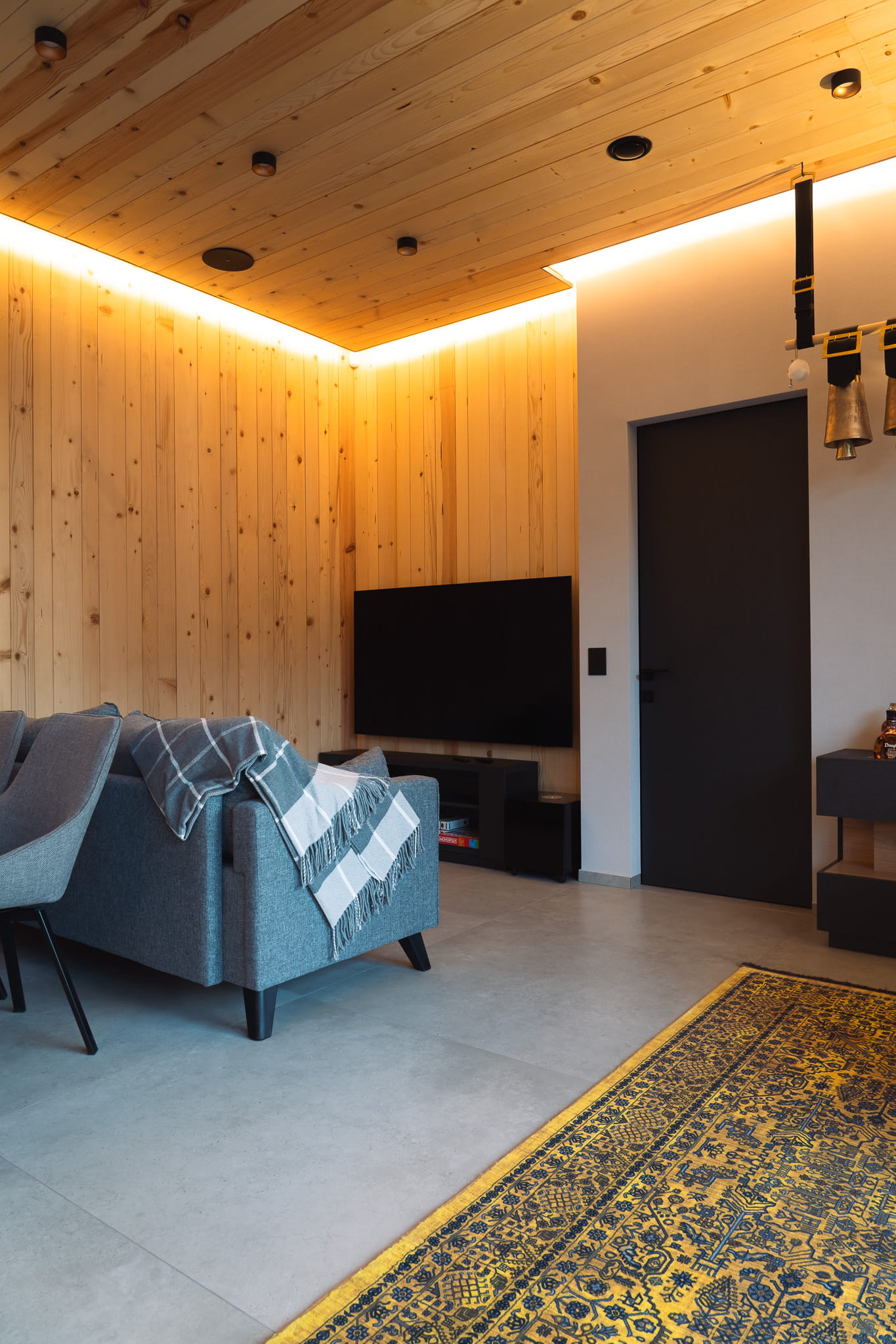 Modern living room with wooden walls, gray sofa with throw blanket, mounted TV, and patterned rug.