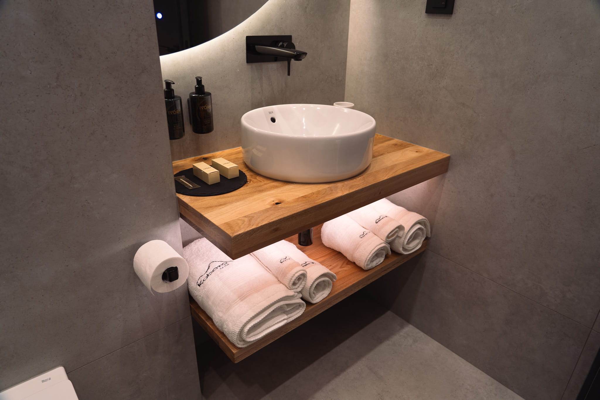 Modern bathroom sink with wooden countertop, white rolled towels below, and toiletries on the side.