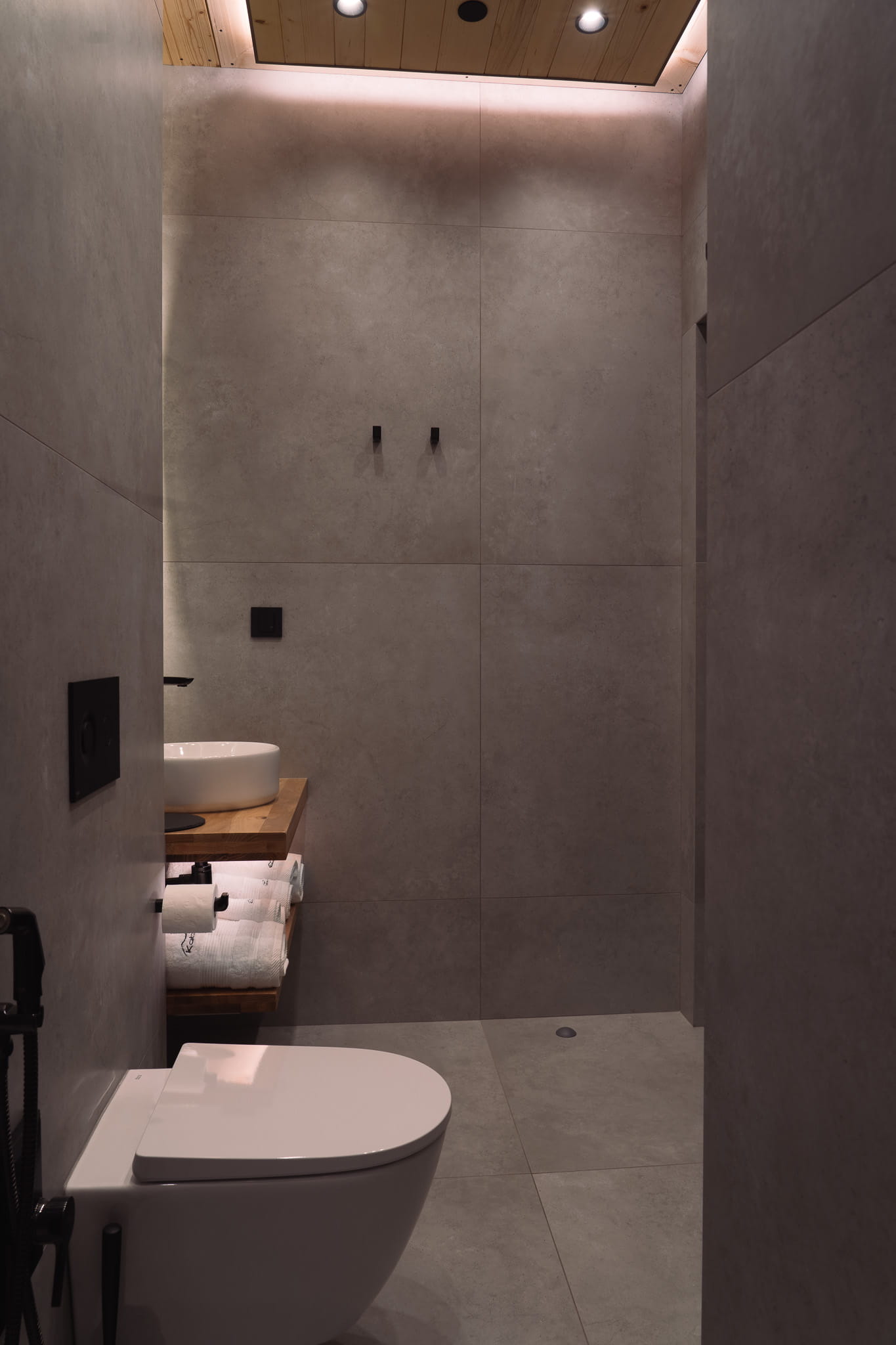 Minimalist bathroom with gray tiled walls, white toilet, wooden sink shelf, and soft ceiling lighting.