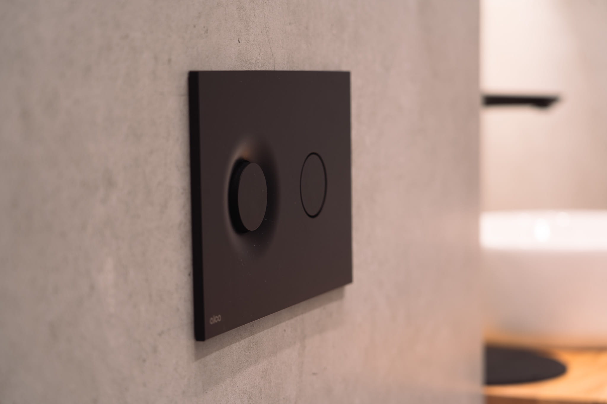 Modern black dual flush toilet button panel mounted on a grey tiled wall.