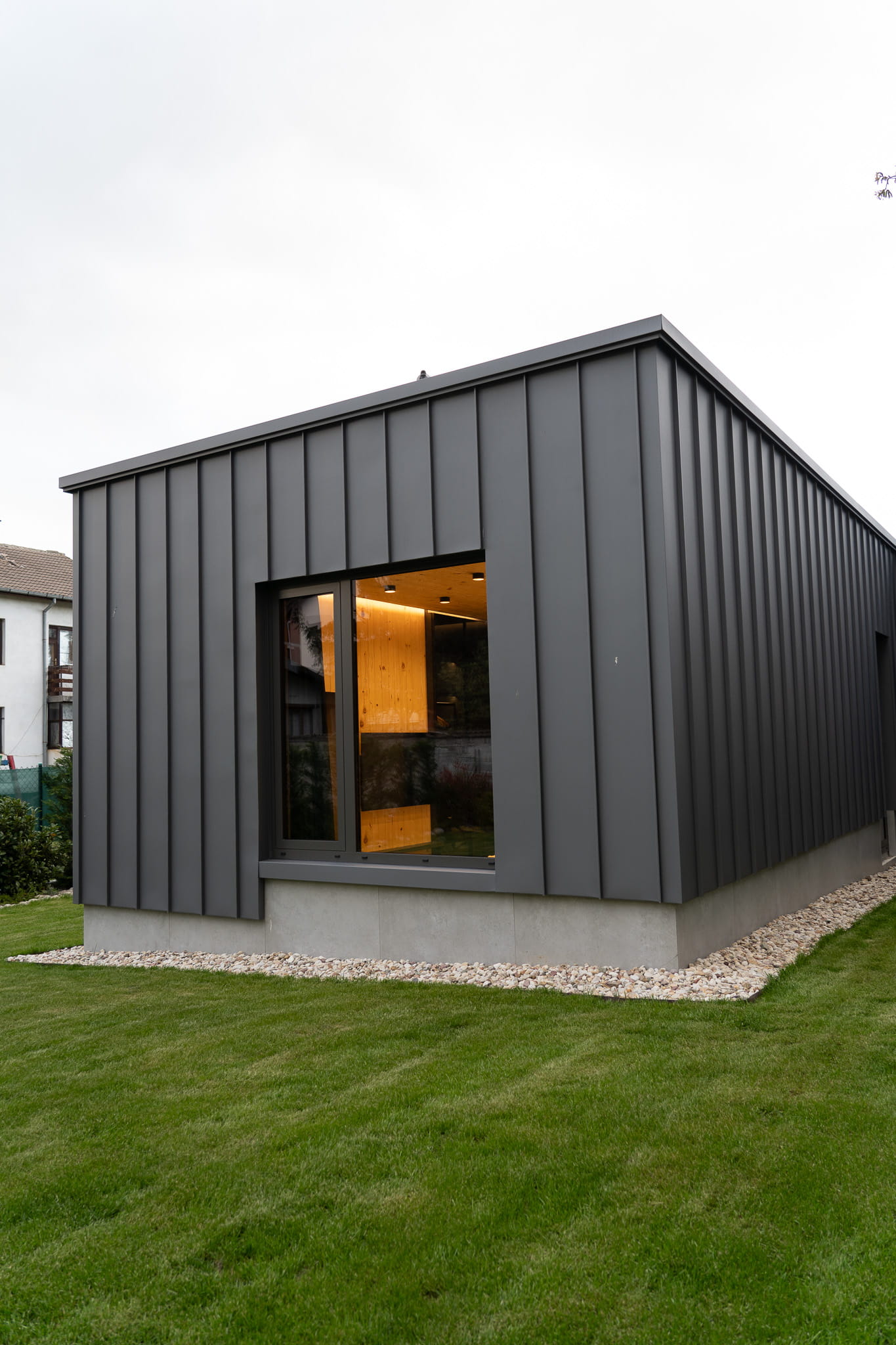 Modern gray metal exterior house with large window showing warm wooden interior and green lawn.