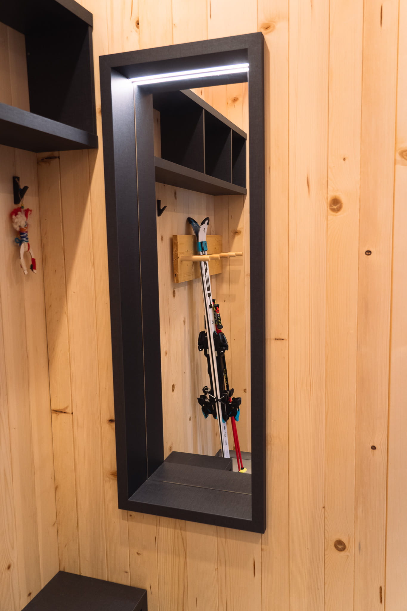 Black-framed mirror on a wooden wall reflecting skis and a wall rack in a cozy room.