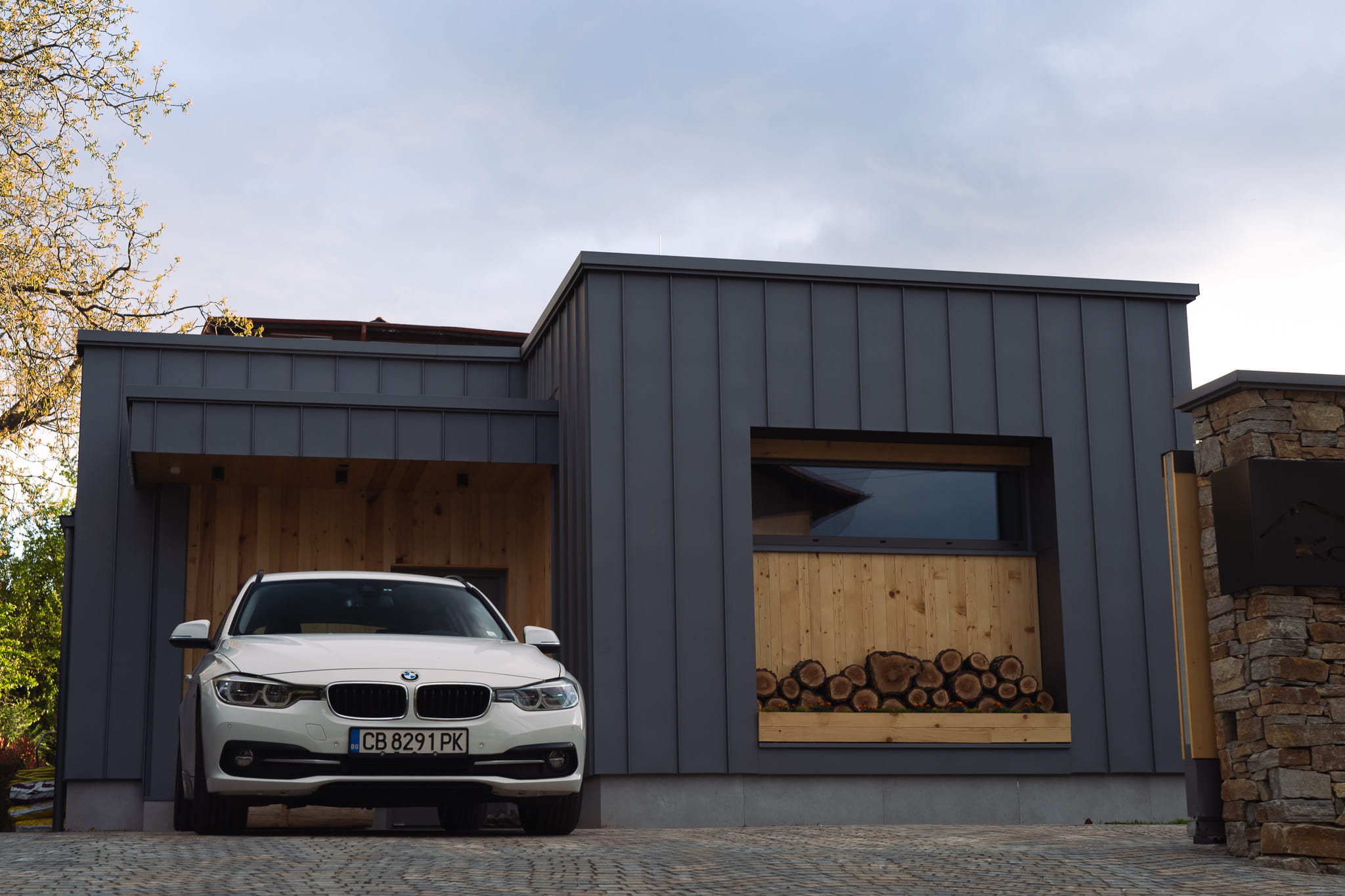 White car parked in front of a modern gray house with wooden accents and stacked firewood.