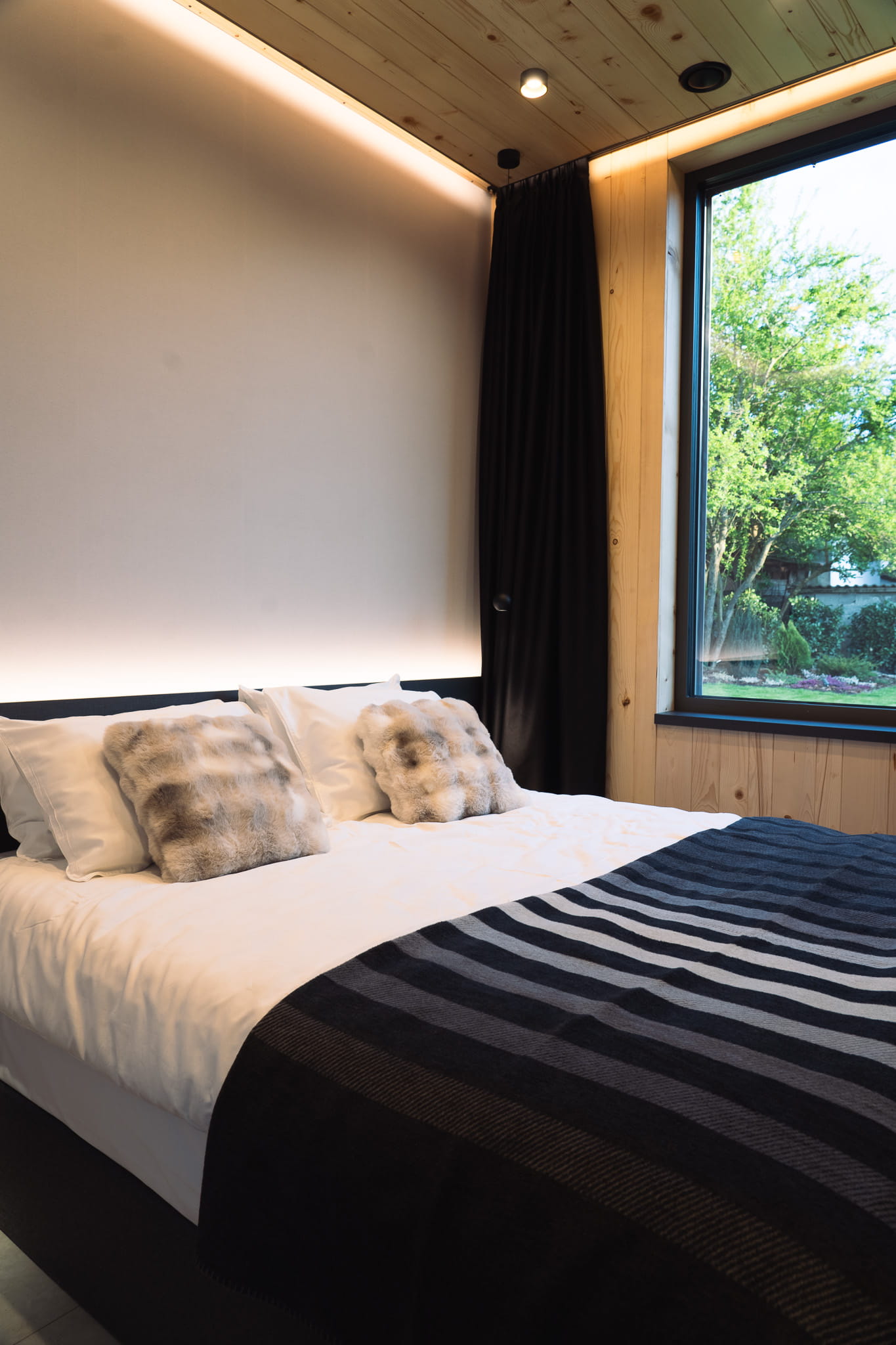 Modern bedroom with a large bed, white linens, fur pillows, a dark striped blanket, and a window showing green trees.