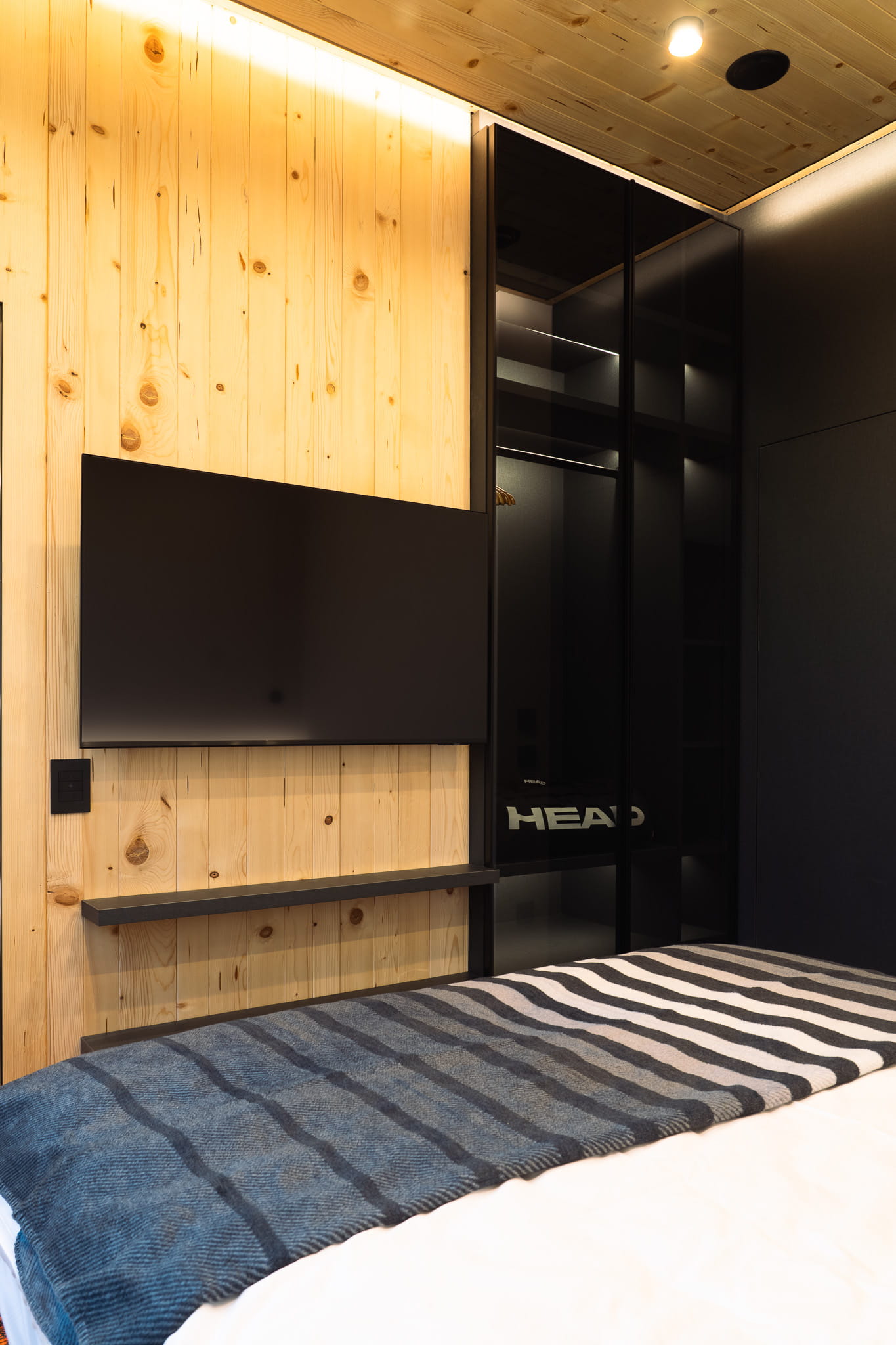 Modern bedroom with wooden paneled wall, black mounted TV, glass door wardrobe, and striped blanket on bed.