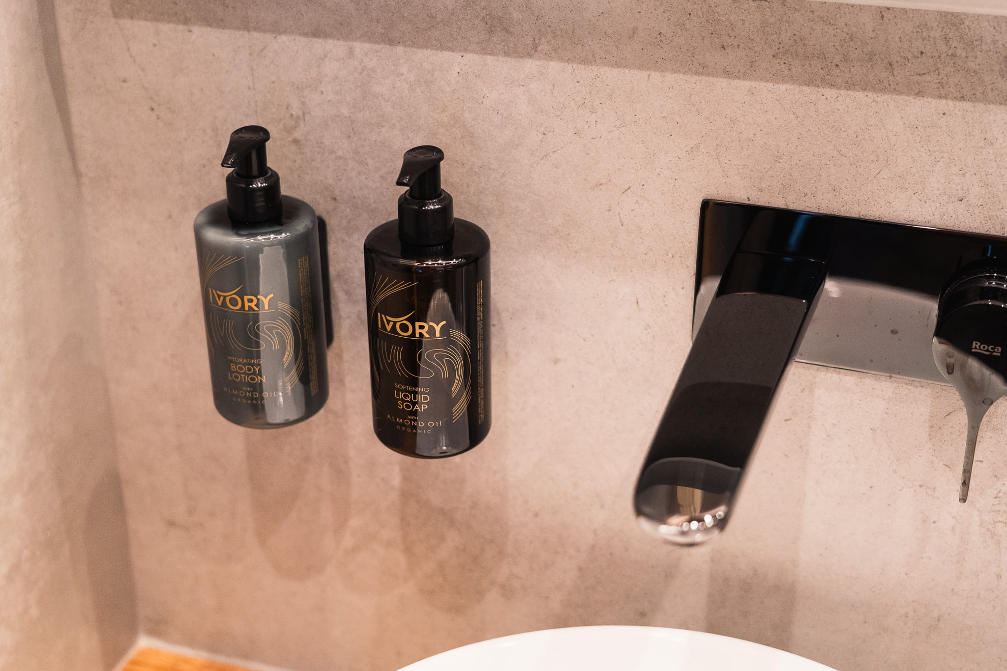 Two pump bottles, one body lotion and one liquid soap, mounted on a bathroom wall next to a modern faucet.