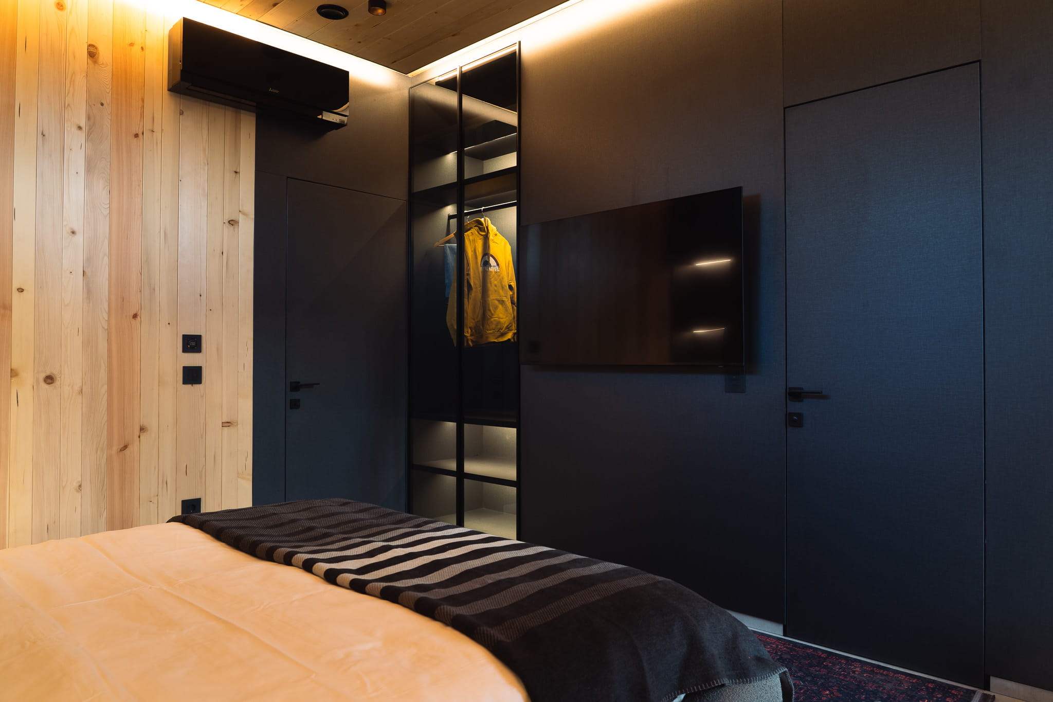 Modern bedroom corner with black walls, wall-mounted TV, glass wardrobe with a yellow jacket, and wood paneling.
