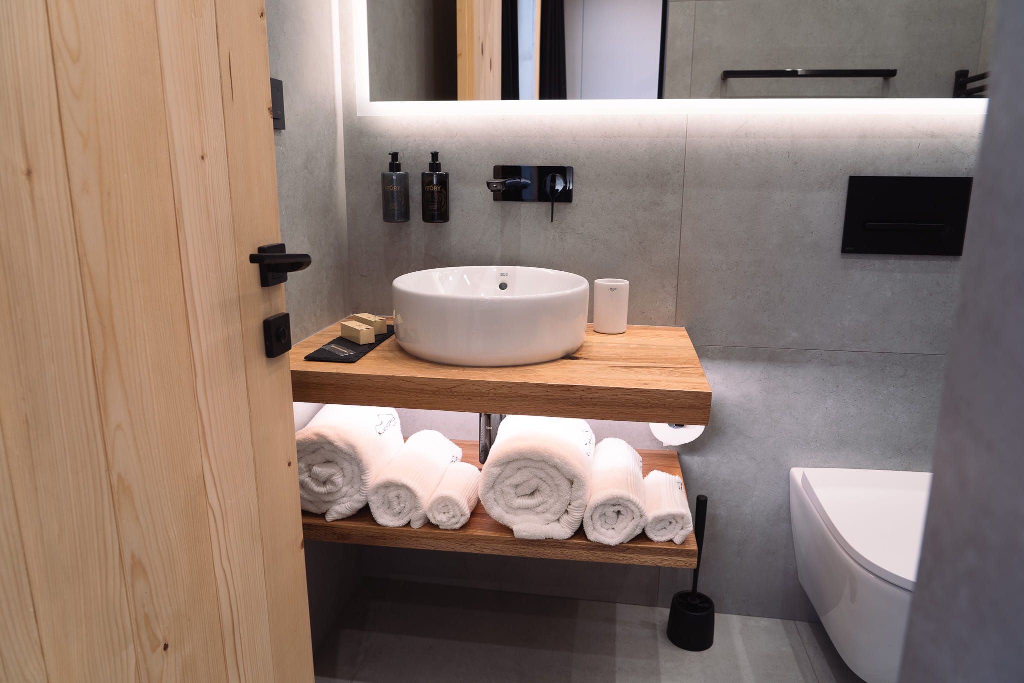 Modern bathroom sink with wooden shelves holding rolled white towels and wall-mounted soap dispensers.