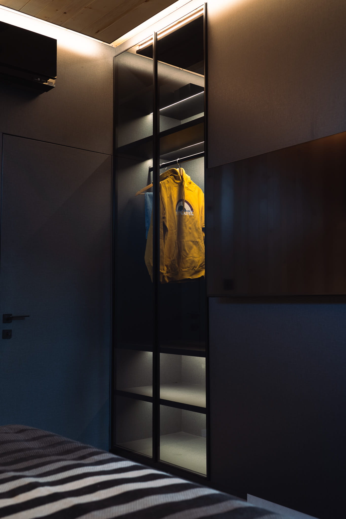 Modern dark closet with glass doors, showing a yellow hoodie hanging inside under soft lighting.
